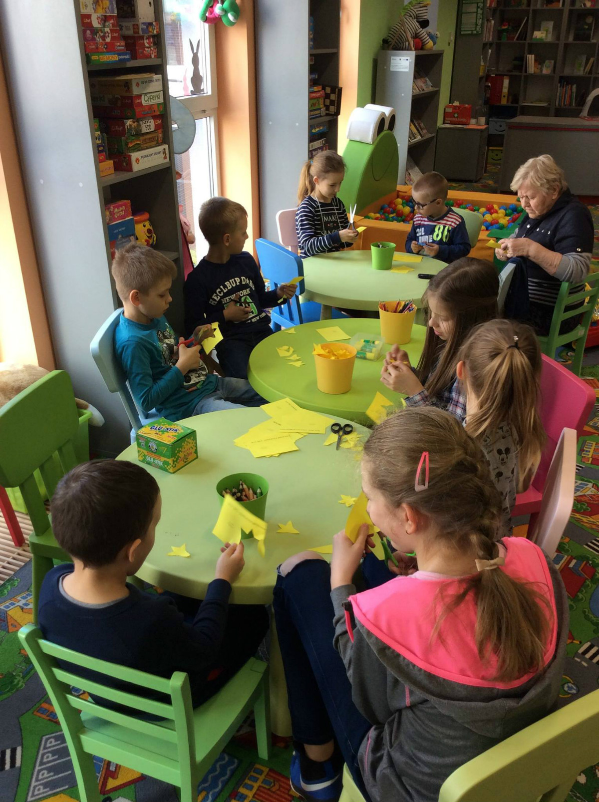 Przy stolikach siedzą dzieci wycinają z żółtego papieru żonkile. 
