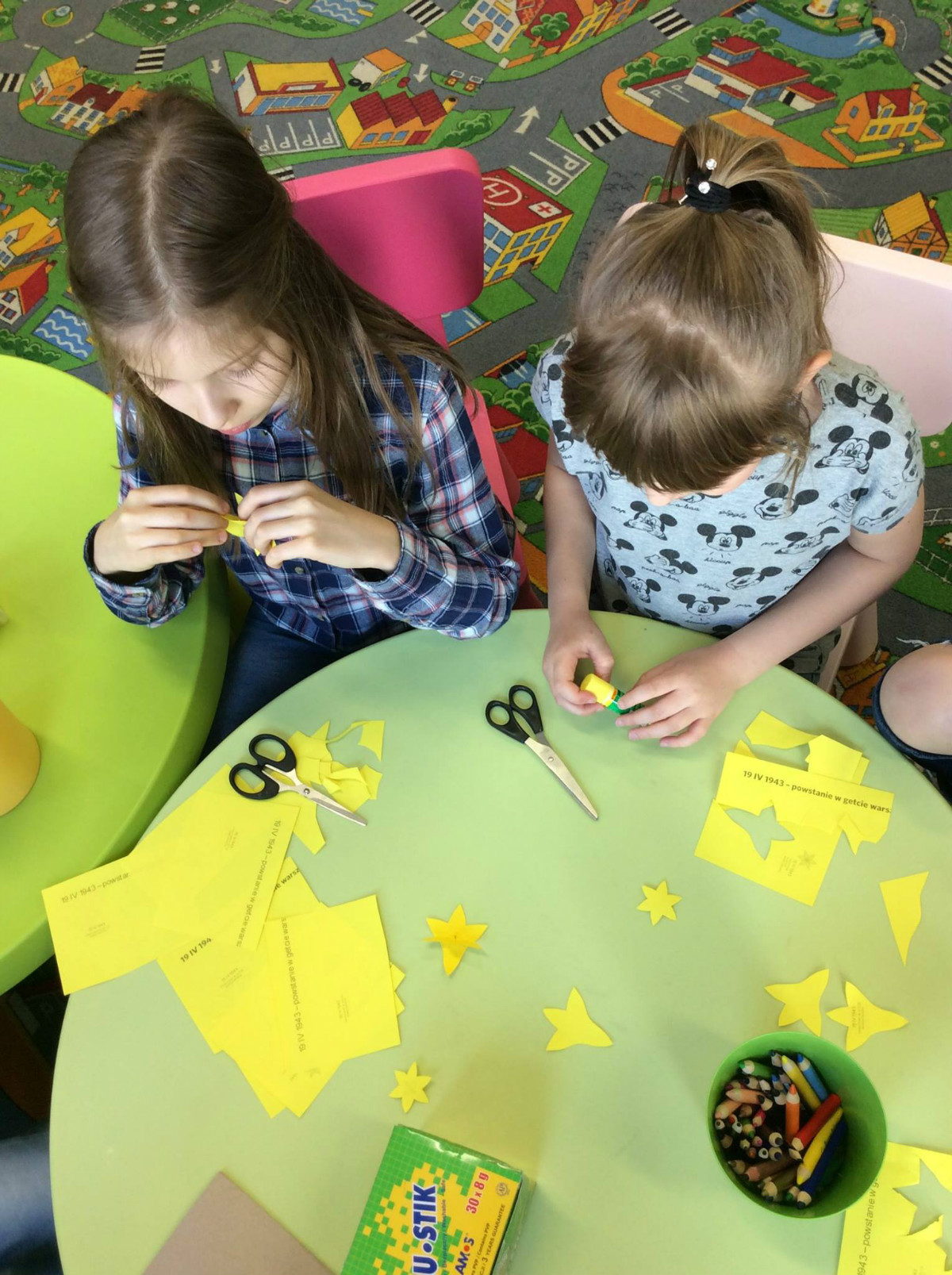 Przy stoliku siedzą dzieci wycinają z żółtego papieru żonkile. 