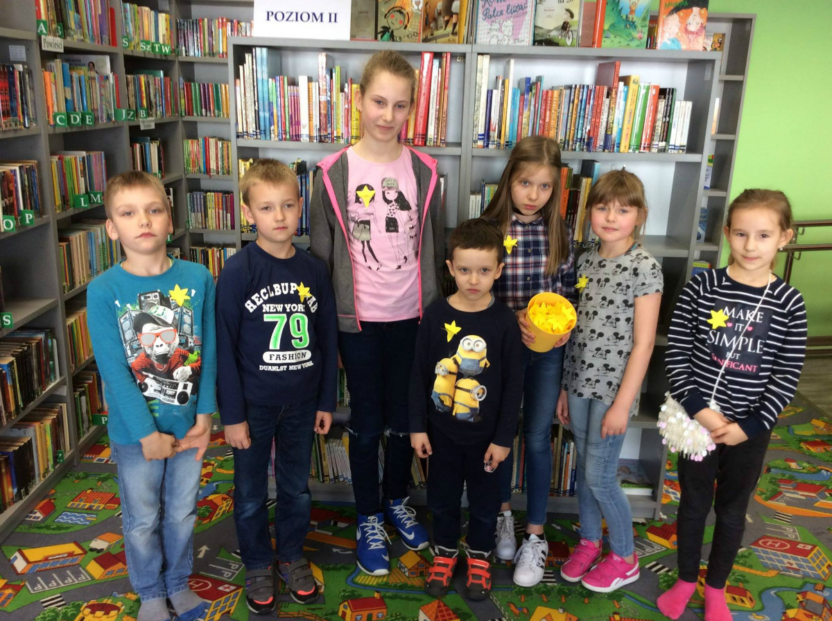 Grupa dzieci  z żółtymi żonkilami stoi przy regale z książkami.
