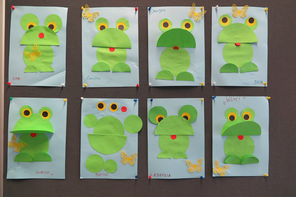 Na tablicy wisi osiem zielonych żabek wykonanych z papieru przez dzieci.