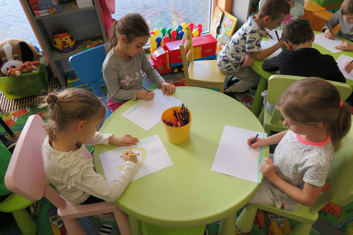 Przy stoliku siedzą dzieci razem rysują kolorowymi kredkami. 