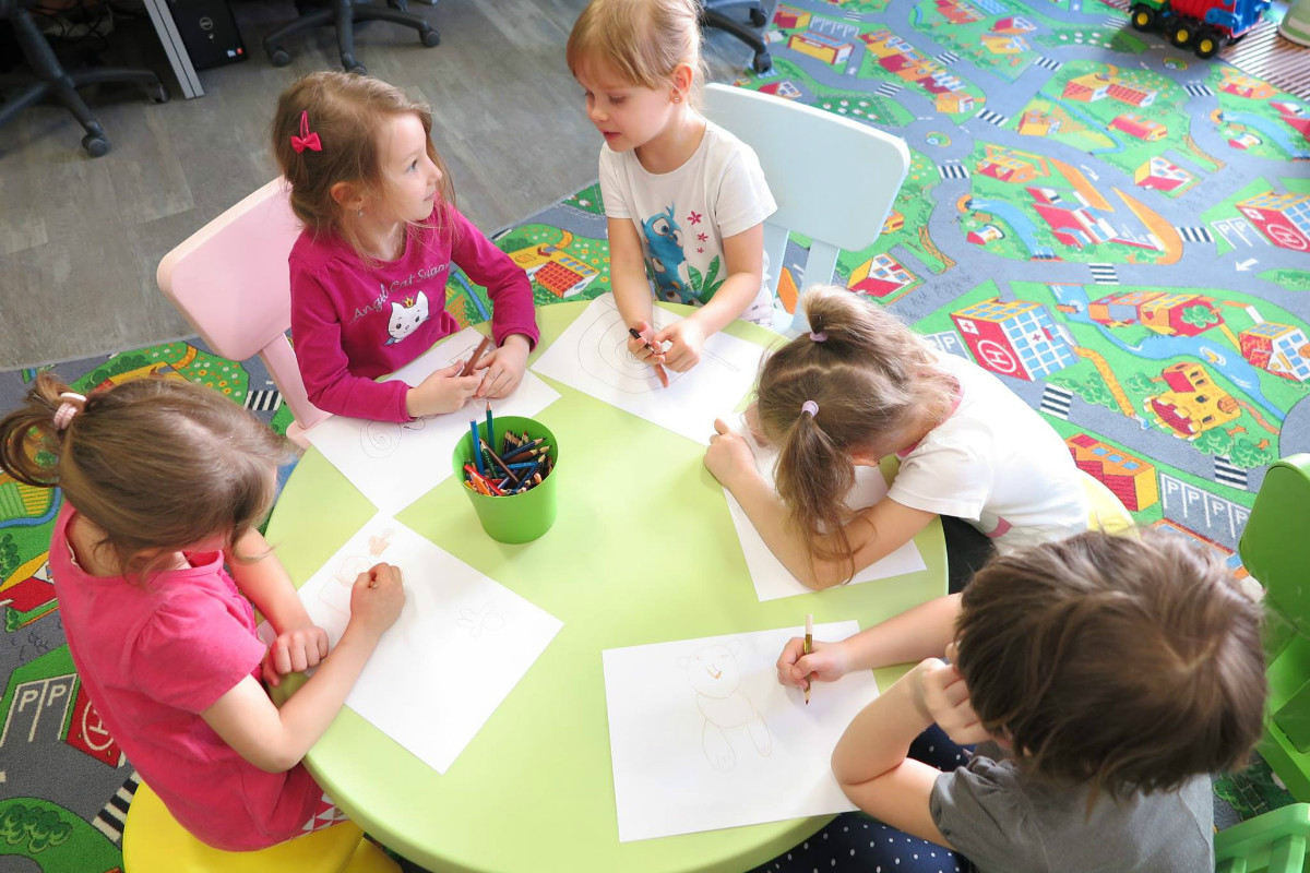 Przy stoliku siedzą dzieci razem rysują kolorowymi kredkami. 