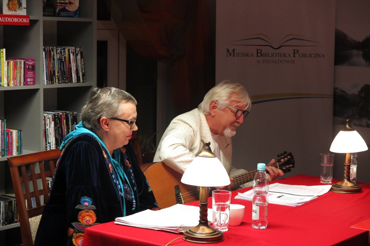 Na scenie przy czerwonym stoliku siedzą aktorzy Wiesława Szymańska i Marian Czarkowski z teatru  im. Stefana Jaracza w Olsztynie. 
