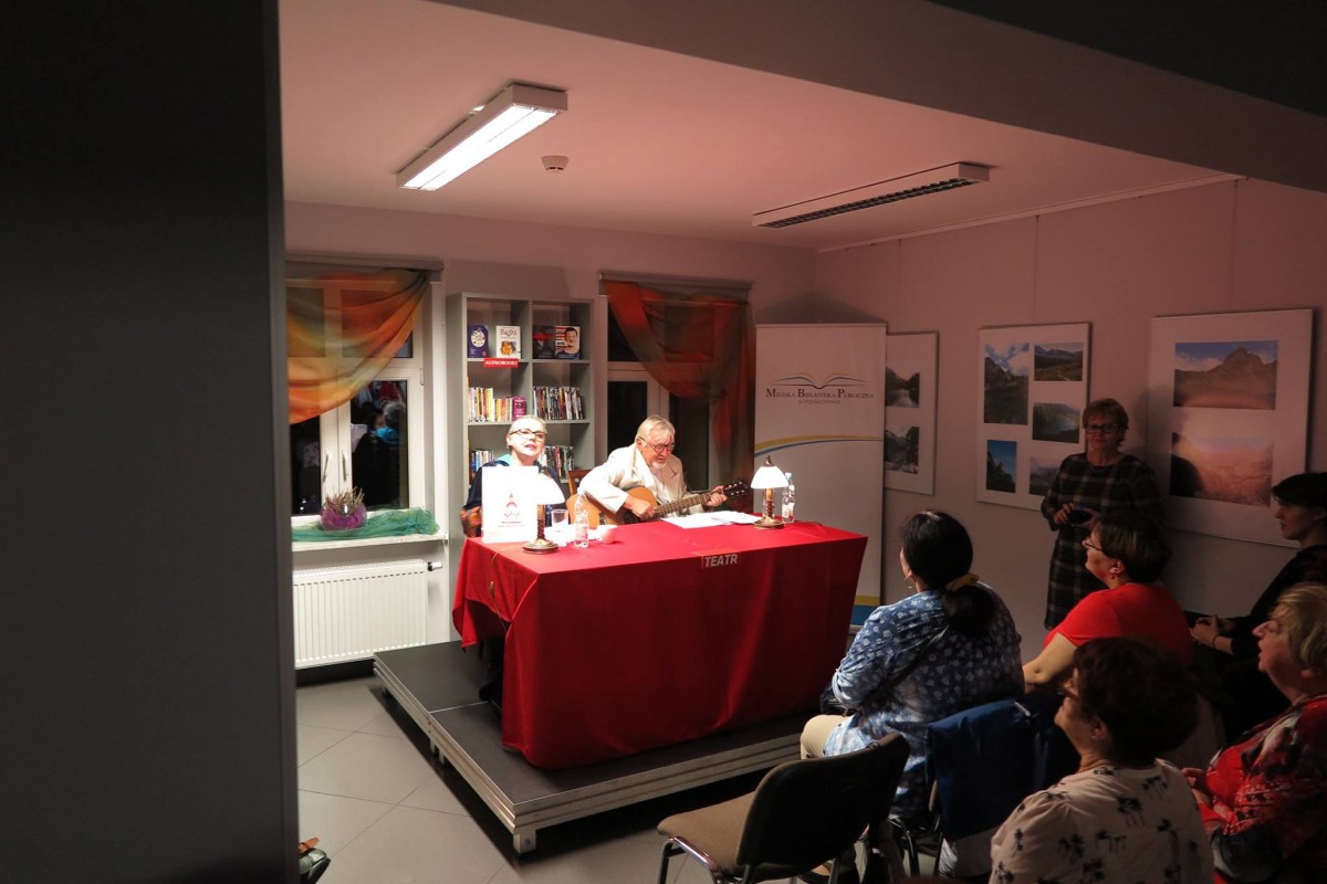 Na scenie przy czerwonym stoliku siedzą aktorzy Wiesława Szymańska i Marian Czarkowski z teatru  im. Stefana Jaracza w Olsztynie obok zgromadzona publiczność.