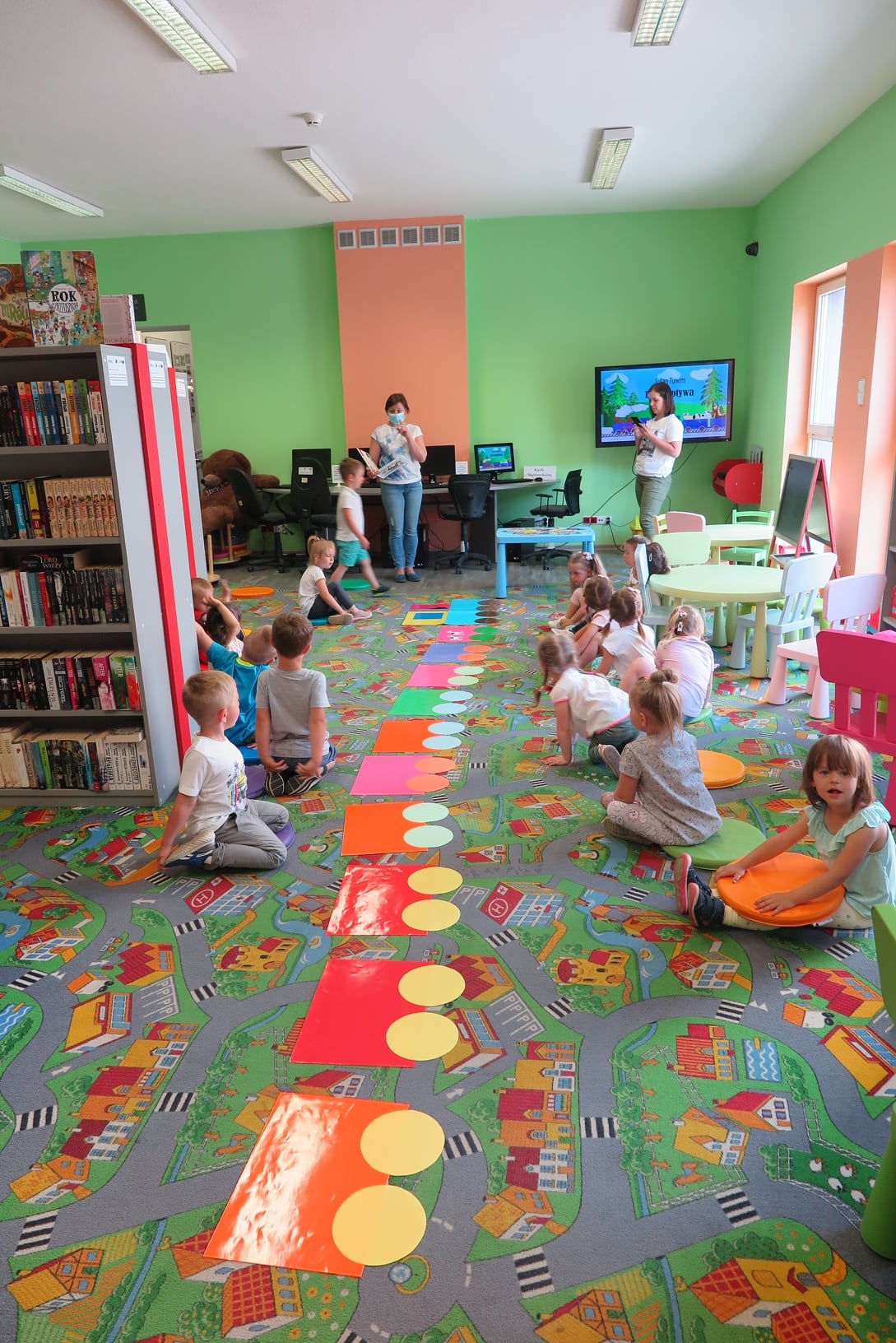 Dzieci siedzą na podłodze w Oddziale dla dzieci w dwóch rzędach. Pomiędzy nimi rozłożone są papierowe kolorowe wagony. Bibliotekarka z książką w ręku stoi przed nimi. Druga kobieta, po prawej stronie robi zdjęcie dzieciom.