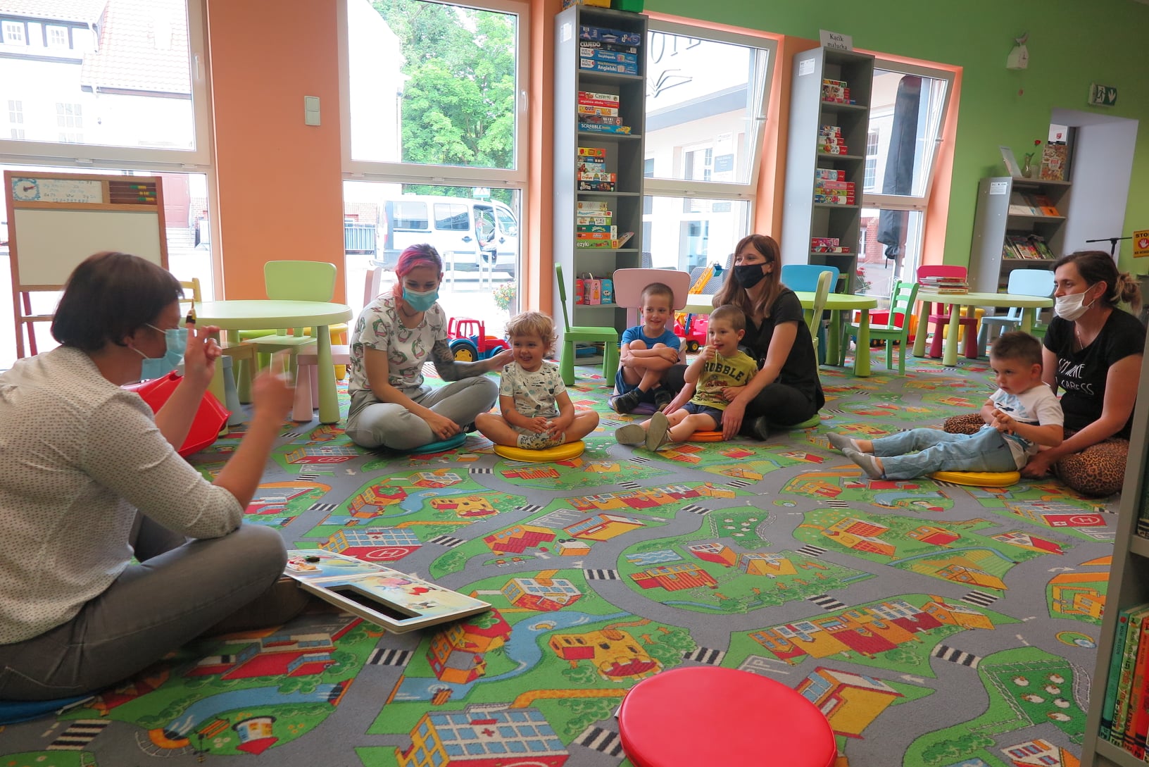 dzieci z rodzicami siedzą na podłodze w oddziale dla dzieci a bibliotekarka czyta im książkę