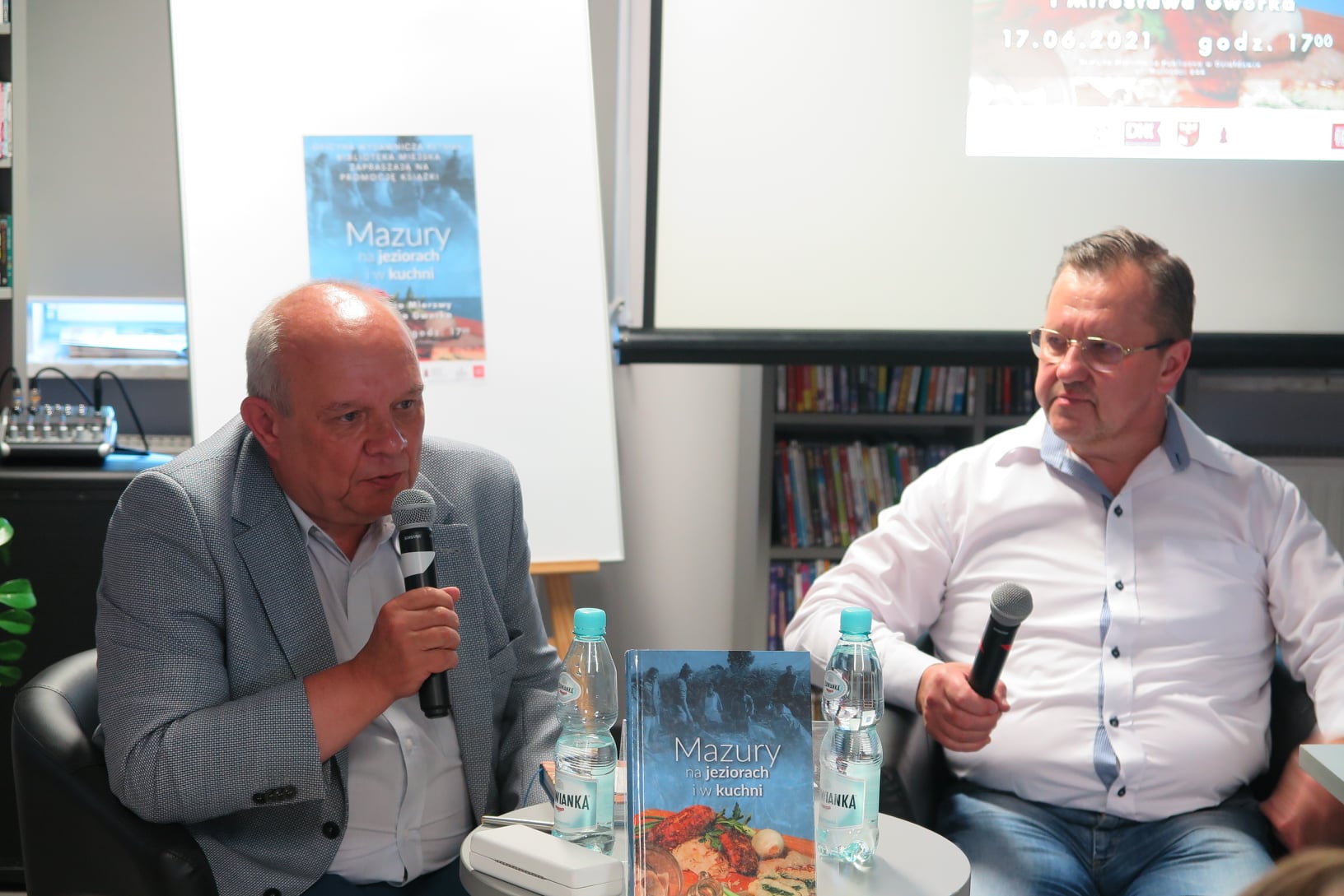 Pomieszczenie biblioteki, przy stoliku siedzą Waldemar Mierzwa i Mirosław Gworek. Waldemar Mierzwa mówi do mikrofonu, który trzyma w dłoni. Na stoliku stoi książka jego autorstwa "Mazury na jeziorach i w kuchni".