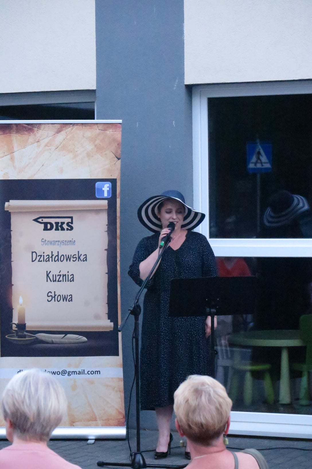 Ubrana w długą czarną suknię i czarno biały kapelusz na głowie kobieta (Marta Wiadrowska), śpiewa przed budynkiem biblioteki dla zebranej publiczności.