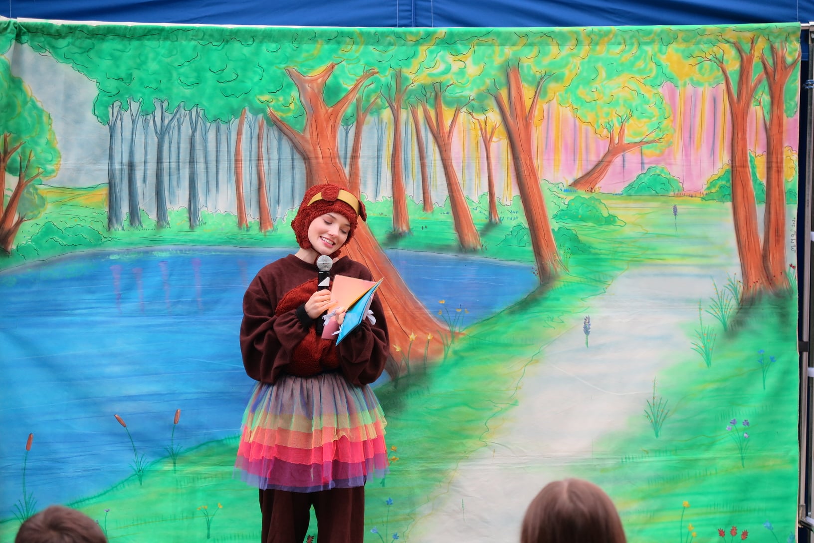 aktorka z teatru Maska przebrana za misia trzyma w ręku mikrofon, stoi na tle dekoracji lasu 