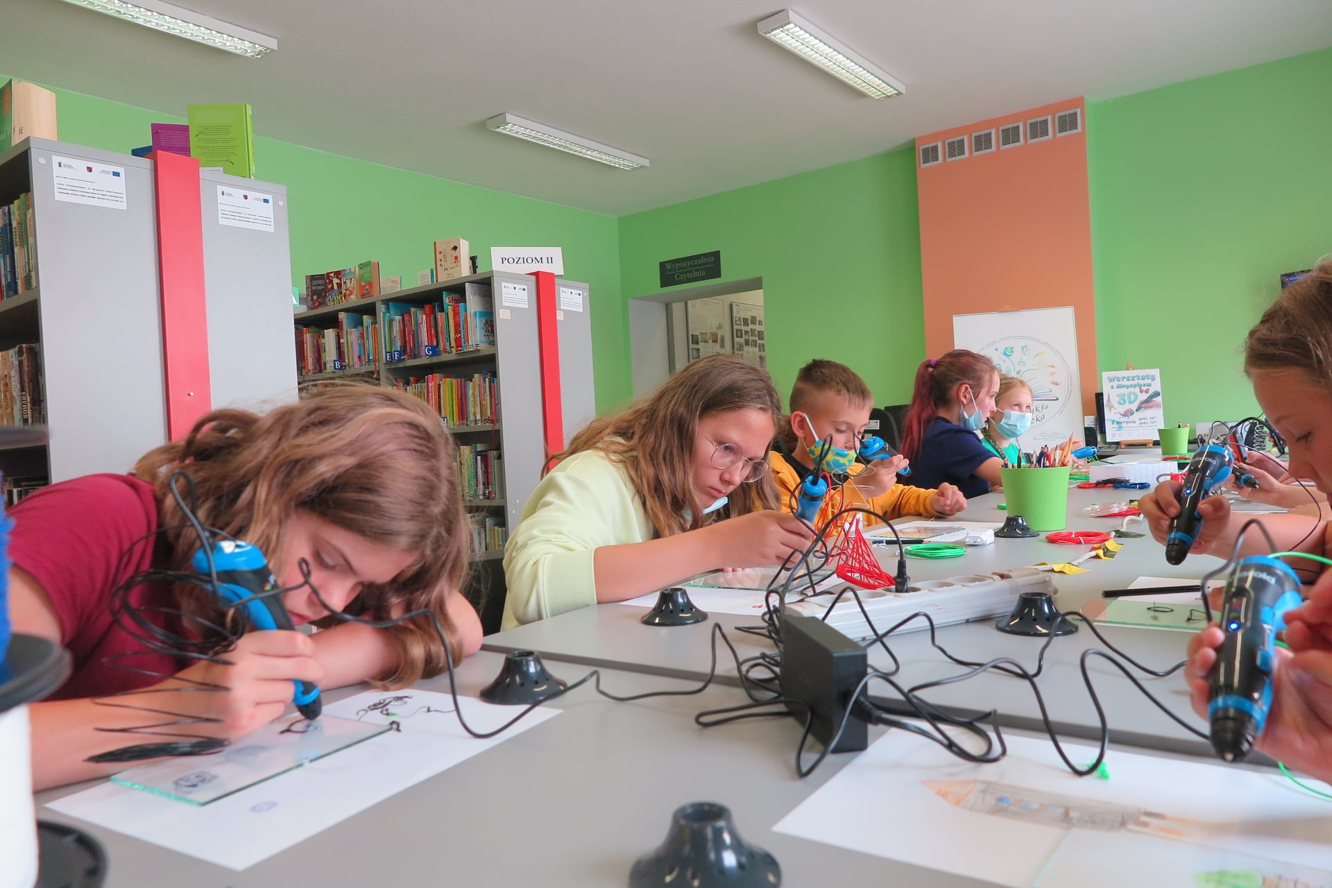 Na zdjęciu grupa dzieci siedząca przy stole w Oddziale dla dzieci. Każde z dzieci trzyma w dłoni długopis 3D. Wykonują nimi wzory na kartkach przykrytymi przezroczystymi szybkami.