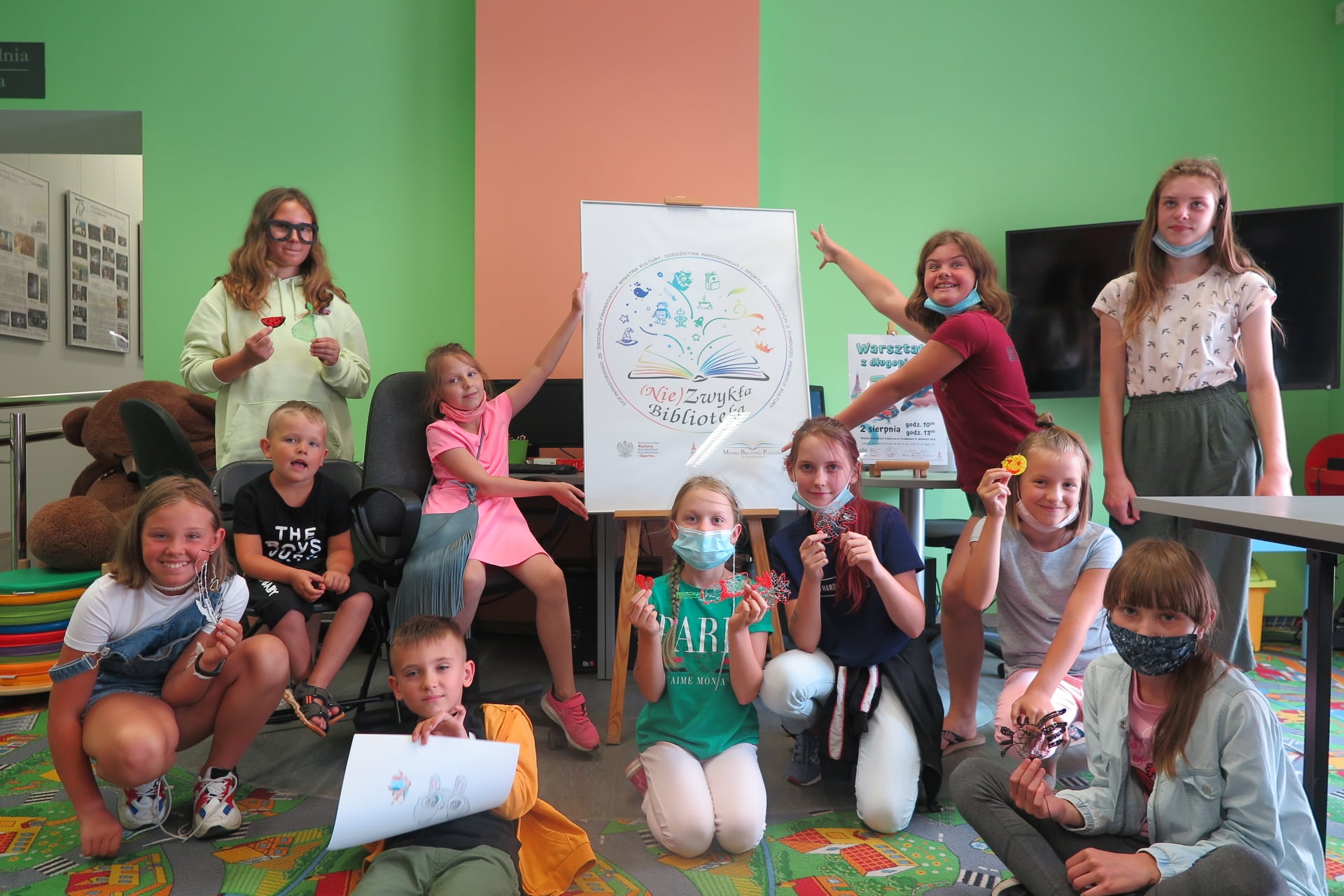 Na zdjęciu grupa dzieci : 9 dziewczynek i 2 chłopców, każde z nich trzyma w dłoni przedmiot wykonany za pomocą długopisu 3D. W tle plakat z logo projektu "(Nie)Zwykła biblioteka".