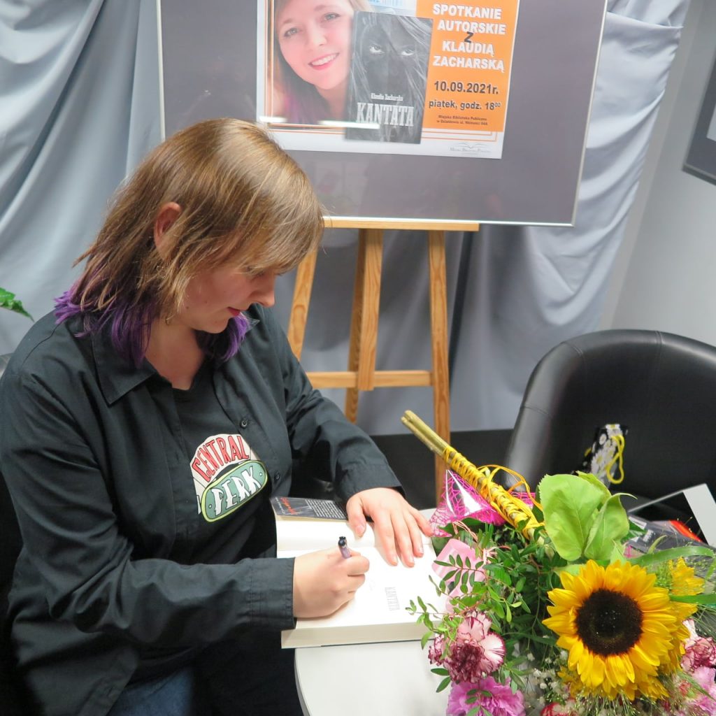 autorka podpisuje książkę siedząc w fotelu, na stoliku leżą kwiaty, w tle plakat promujący spotkanie autorskie