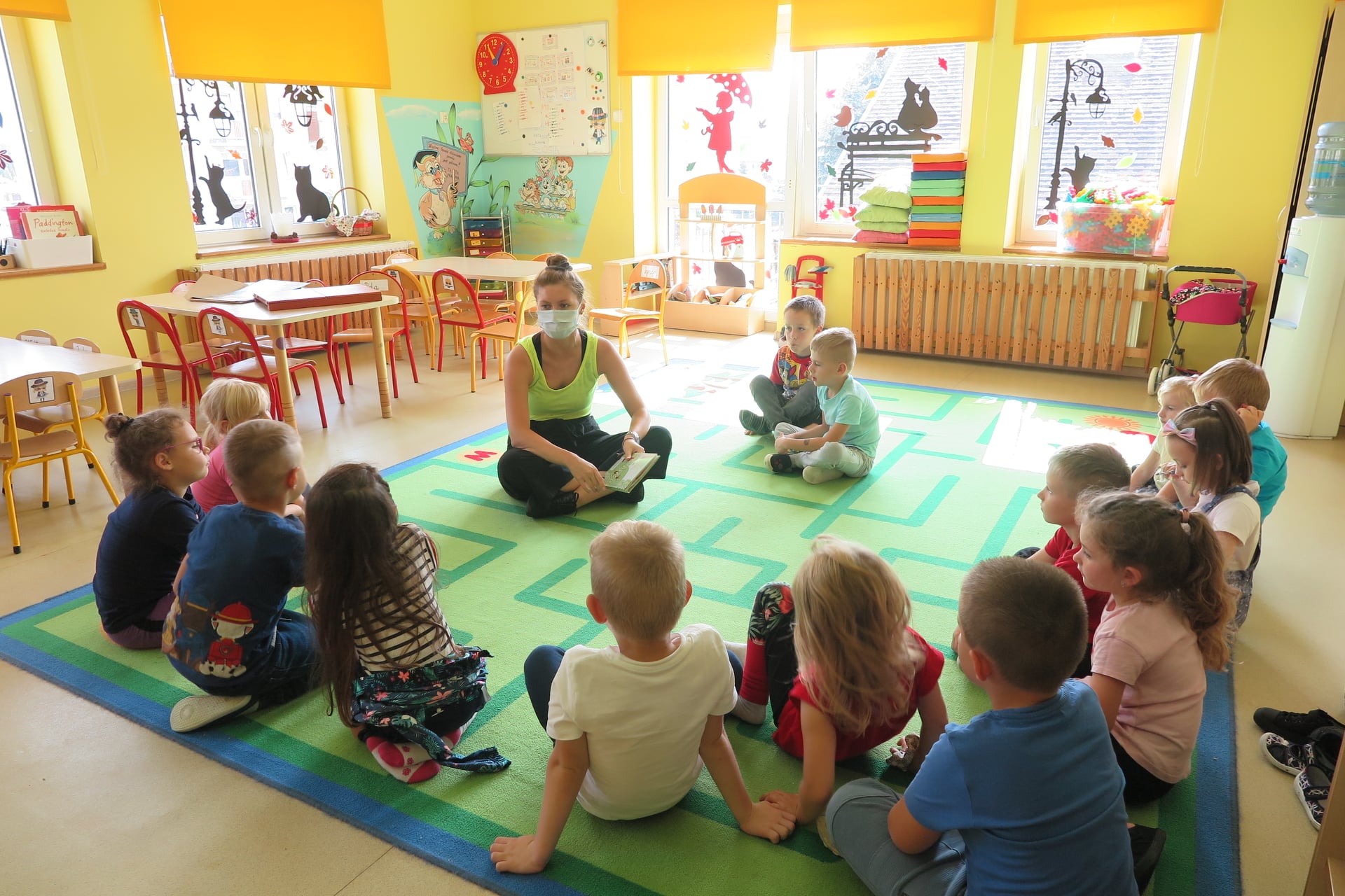 Na zdjęciu grupa dzieci siedząca na dywanie w przedszkolu. Po środku siedzi bibliotekarka, w dłoniach trzyma książkę.