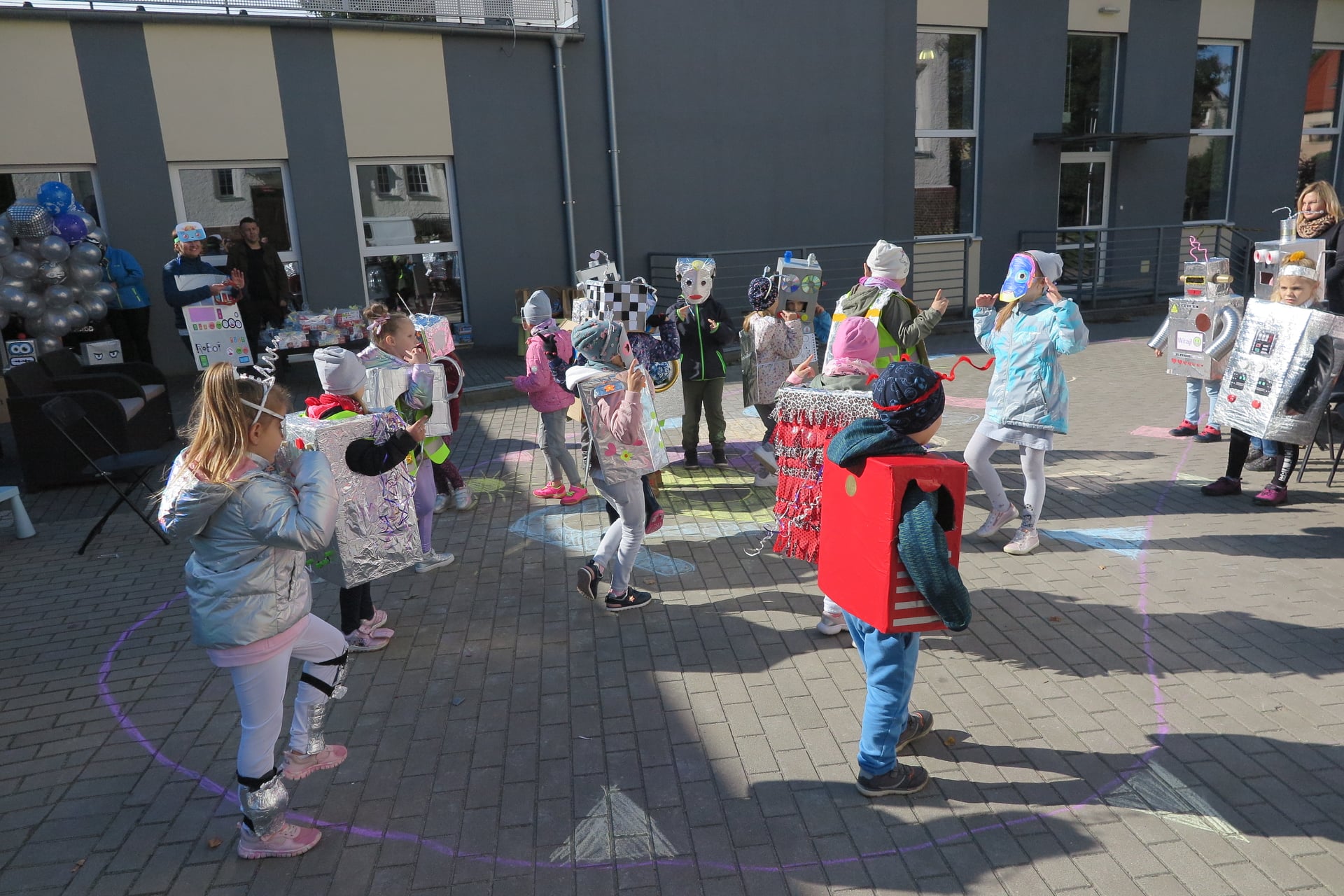 plac przed biblioteką, na placu inna grupa dzieci przebrana za roboty tańczy, w tle widać budynek biblioteki, organizatora marszu i balony