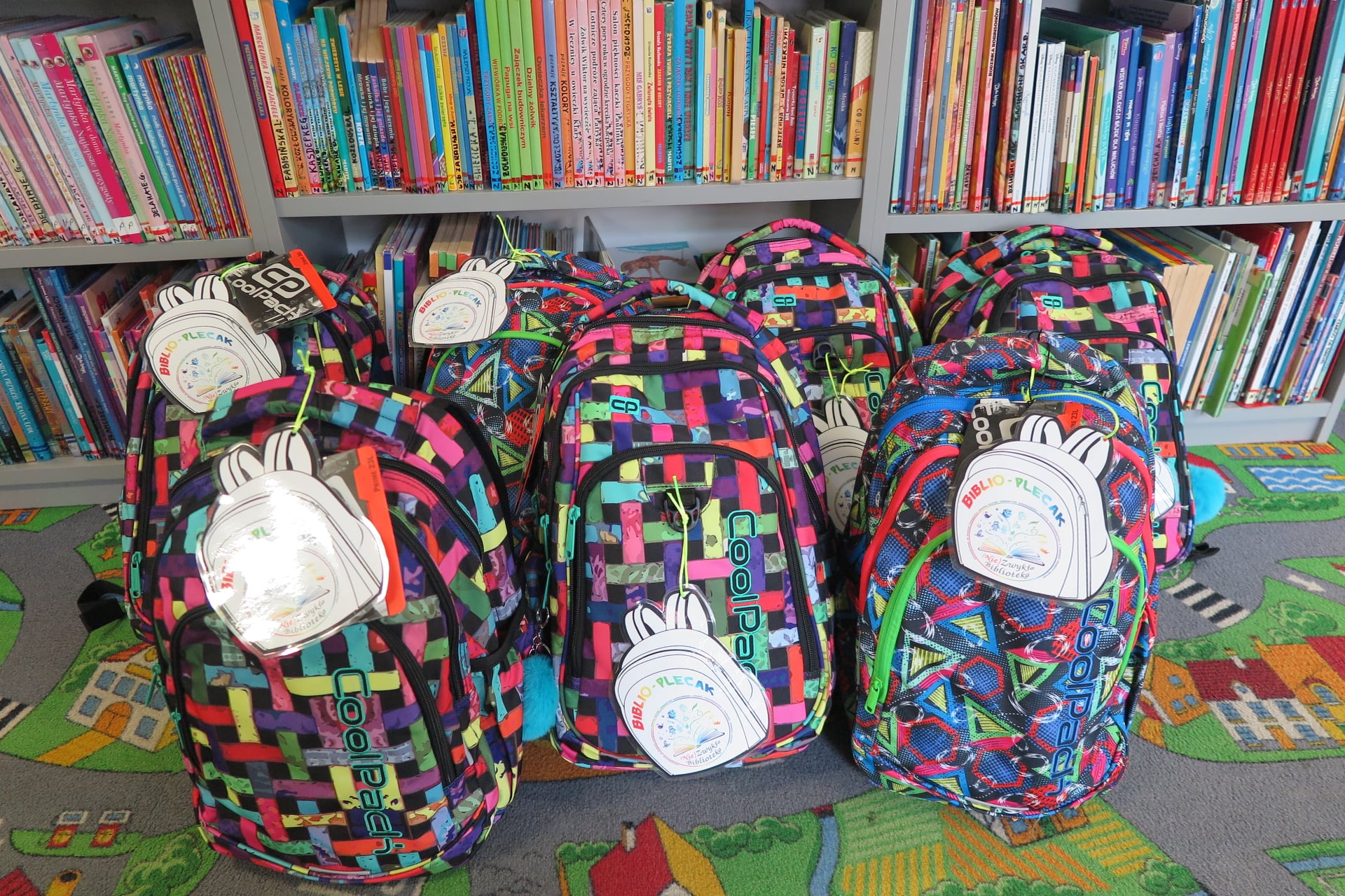 7 kolorowych plecaków leży na podłodze w oddziale dla dzieci oparte o regał z książkami