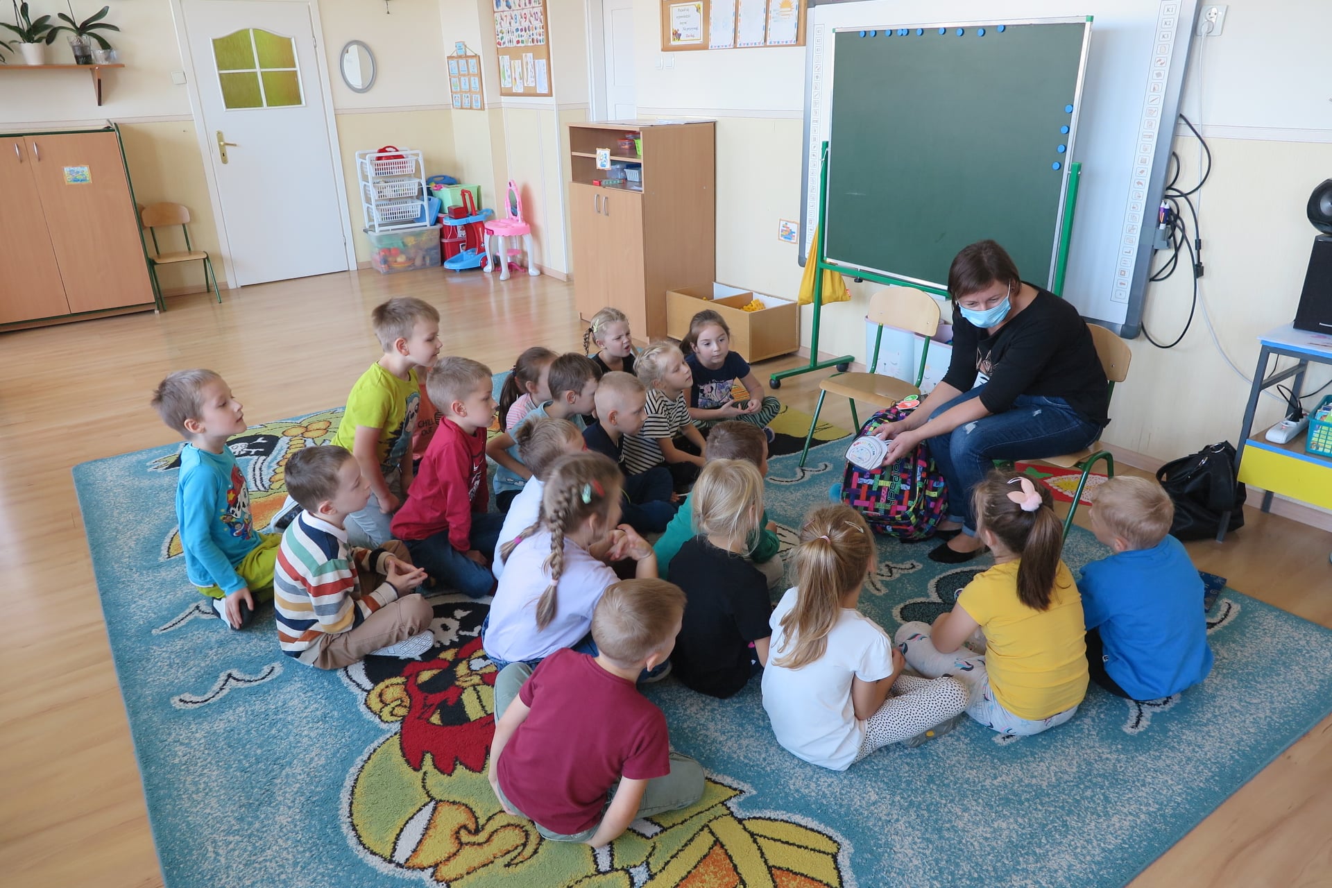 sala w przedszkolu, na dywanie na podłodze podłodze siedzi 17 dzieci, bibliotekarka siedzi na krześle trzymając w rekach biblioplecek