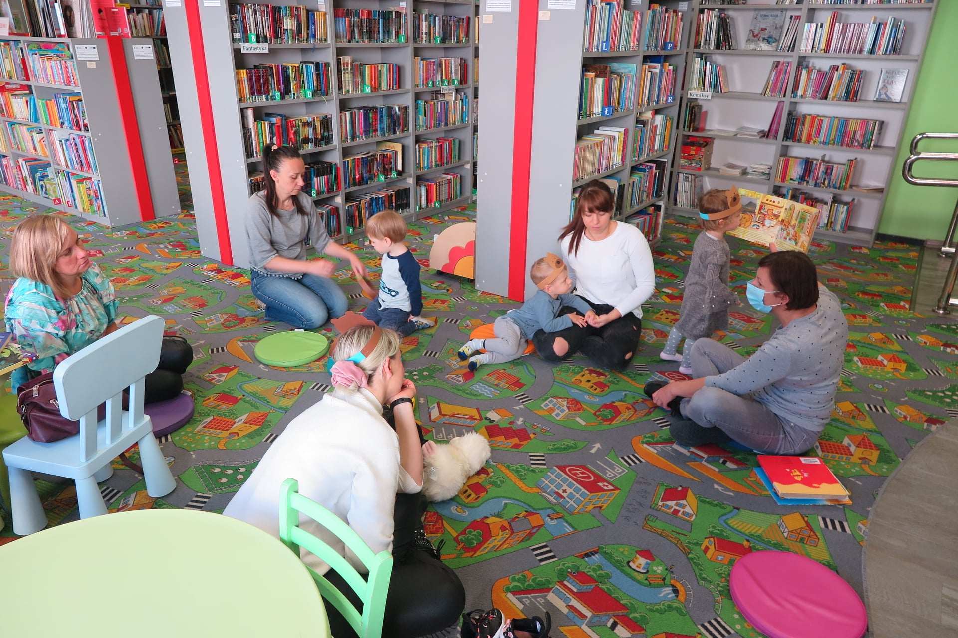 pięć kobiet siedzi na podłodze w oddziale dla dzieci, obok siedzi dwoje dzieci, jedno dziecko stoi i przygląda się obrazków w książce trzymanej przez bibliotekarkę, w tle widać regały z książkami