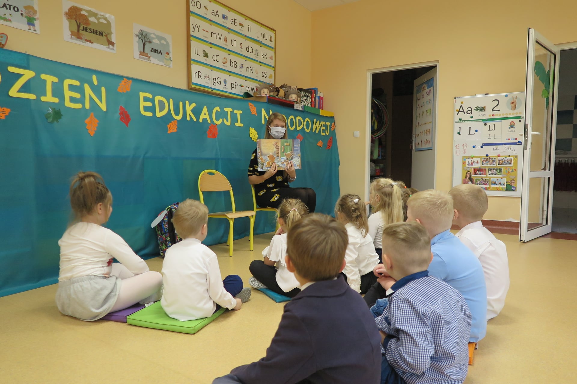 Sala przedszkolna, grupa dzieci siedzi na ziemi dookoła bibliotekarki, która trzyma w dłoni książkę odwróconą obrazkami w stronę dzieci. Bibliotekarka siedzi na krześle na twarzy ma maseczkę ochronną. 