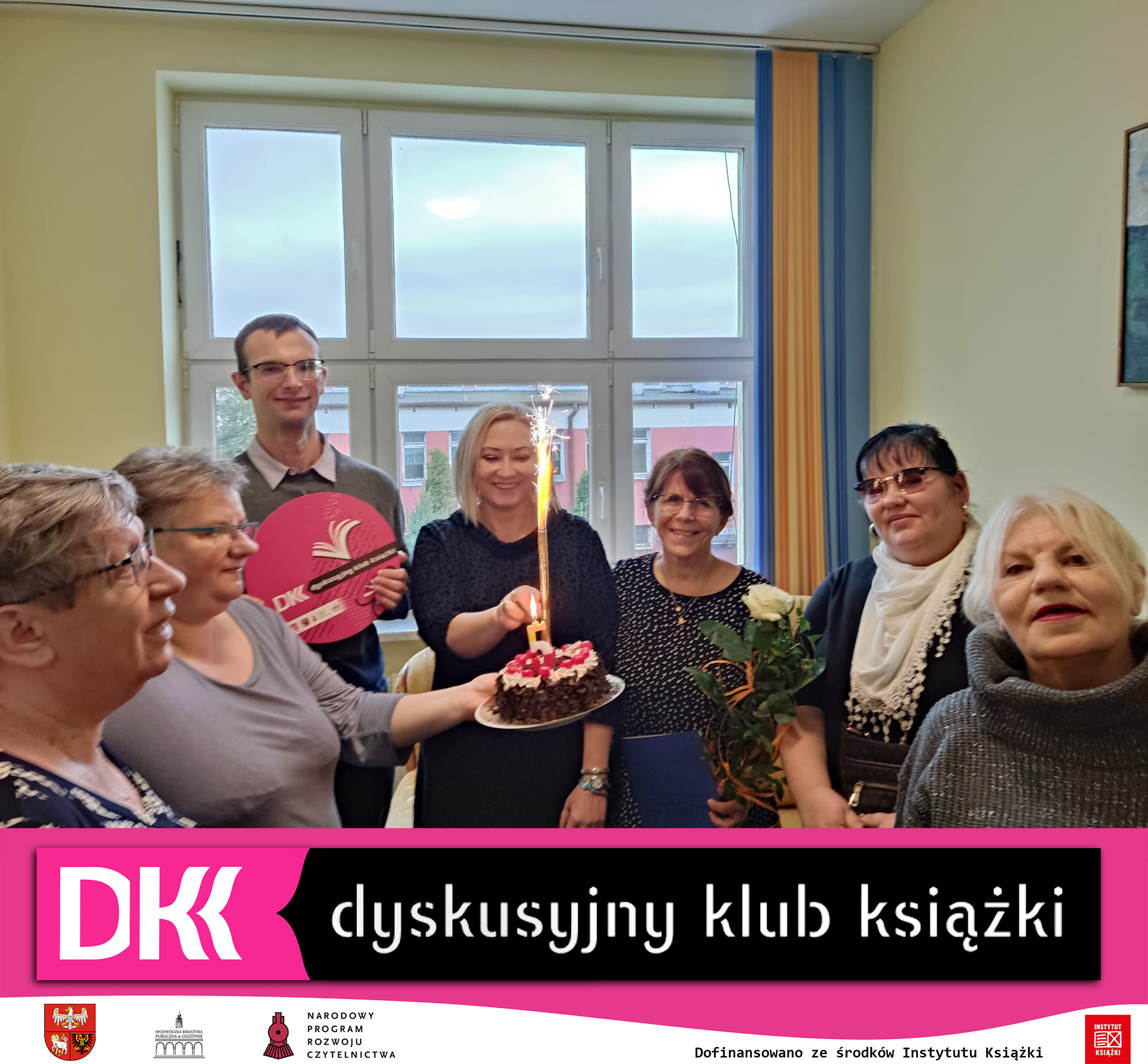 6 kobiet i jeden mężczyzna, jedna z kobiet trzyma w ręku tort ze świeczką w kształcie cyfry 5 i zimnym ogniem, inna kobieta trzyma w ręku kwiaty, na dole zdjęcia widnieje logo klubu DKK