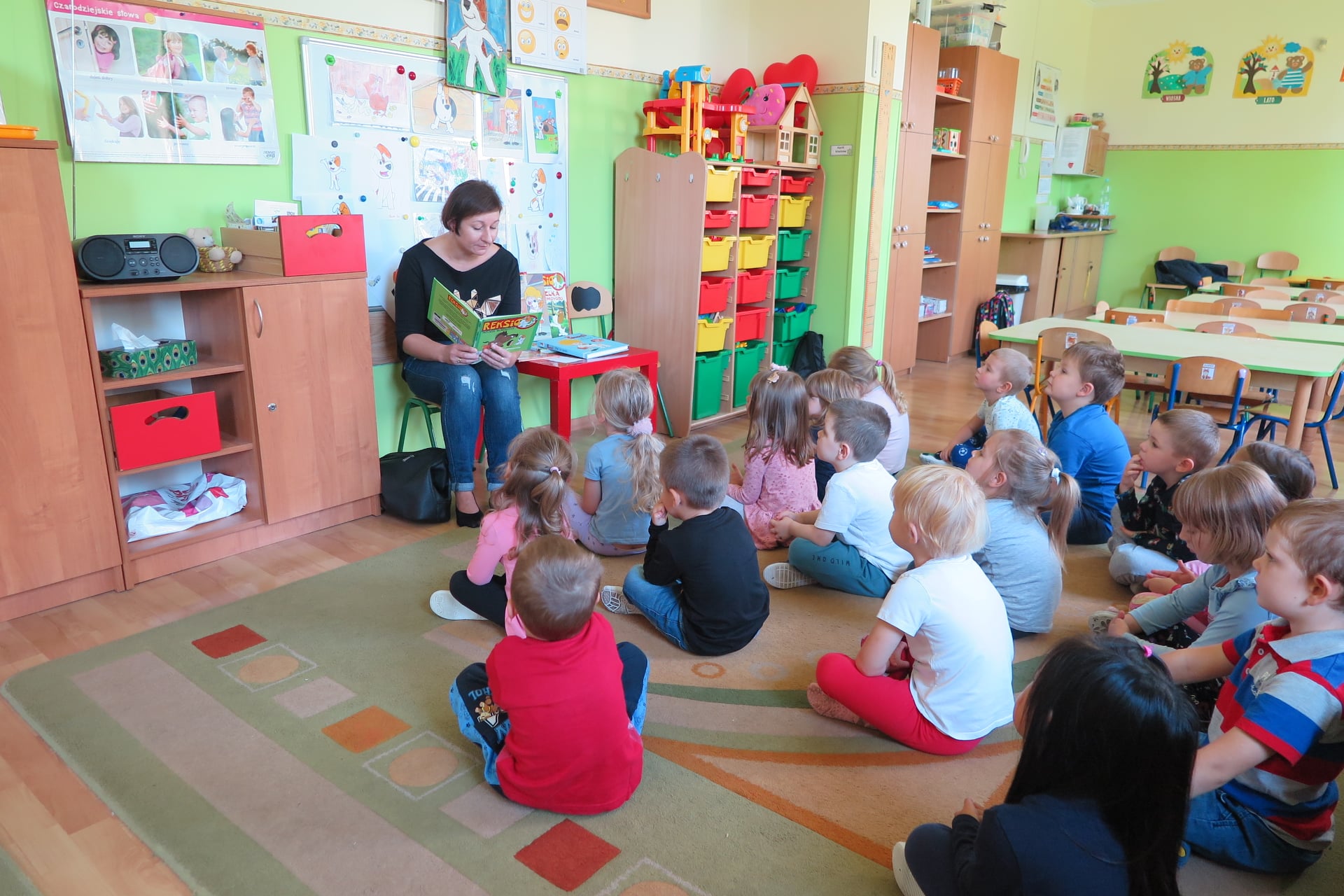 sala w przedszkolu a w niej grupa dzieci siedzi na dywanie zwrócone w stronę bibliotekarki siedzącej na krześle i czytającej książkę
