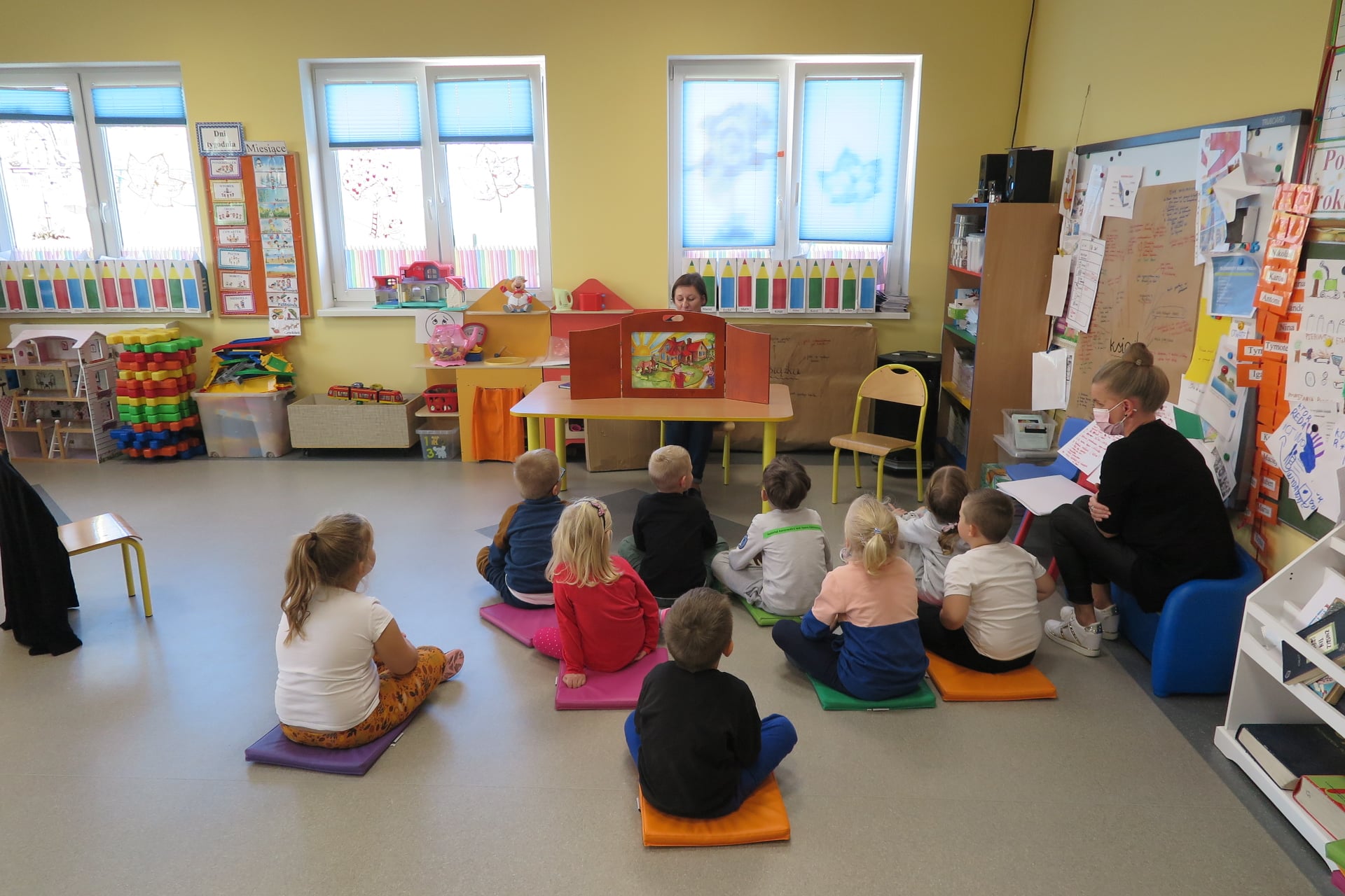 sala w przedszkolu a w niej grupa dzieci siedzi na matach, zwrócone w stronę bibliotekarki siedzącej na krześle przy stole, przed nią rozstawiony jest teatrzyk Kamishibai, obok grupy na krześle dzieci wychowawczyni