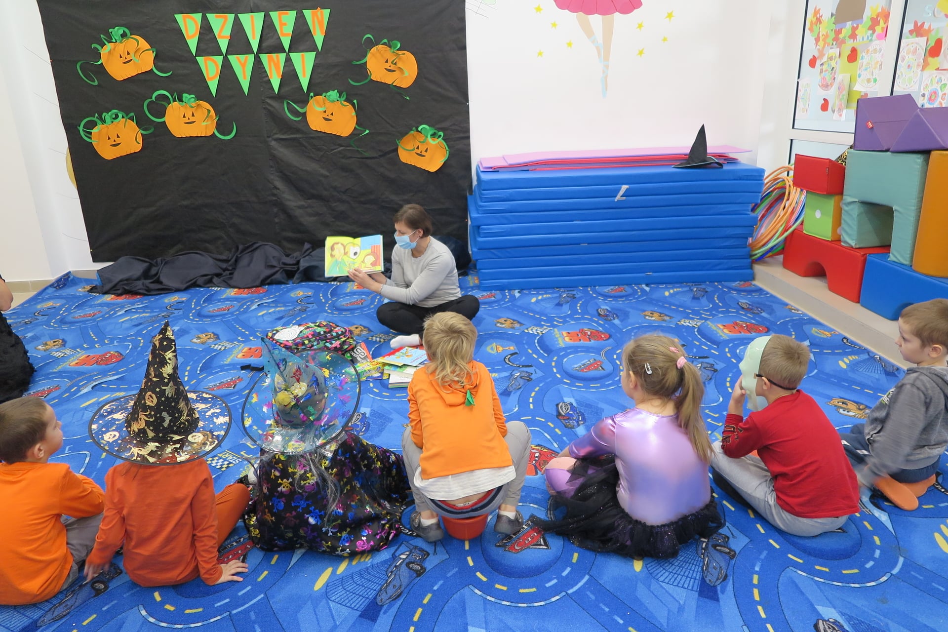 sala przedszkolna udekorowana w klimatach Halloween, 7 dzieci siedzi na dywanie przed bibliotekarką, obok niej leży Biblioplecek z wyłożoną zawartością , niektóre dzieci mają przebrania halloweenowe, w tle widać napis dzień dyni