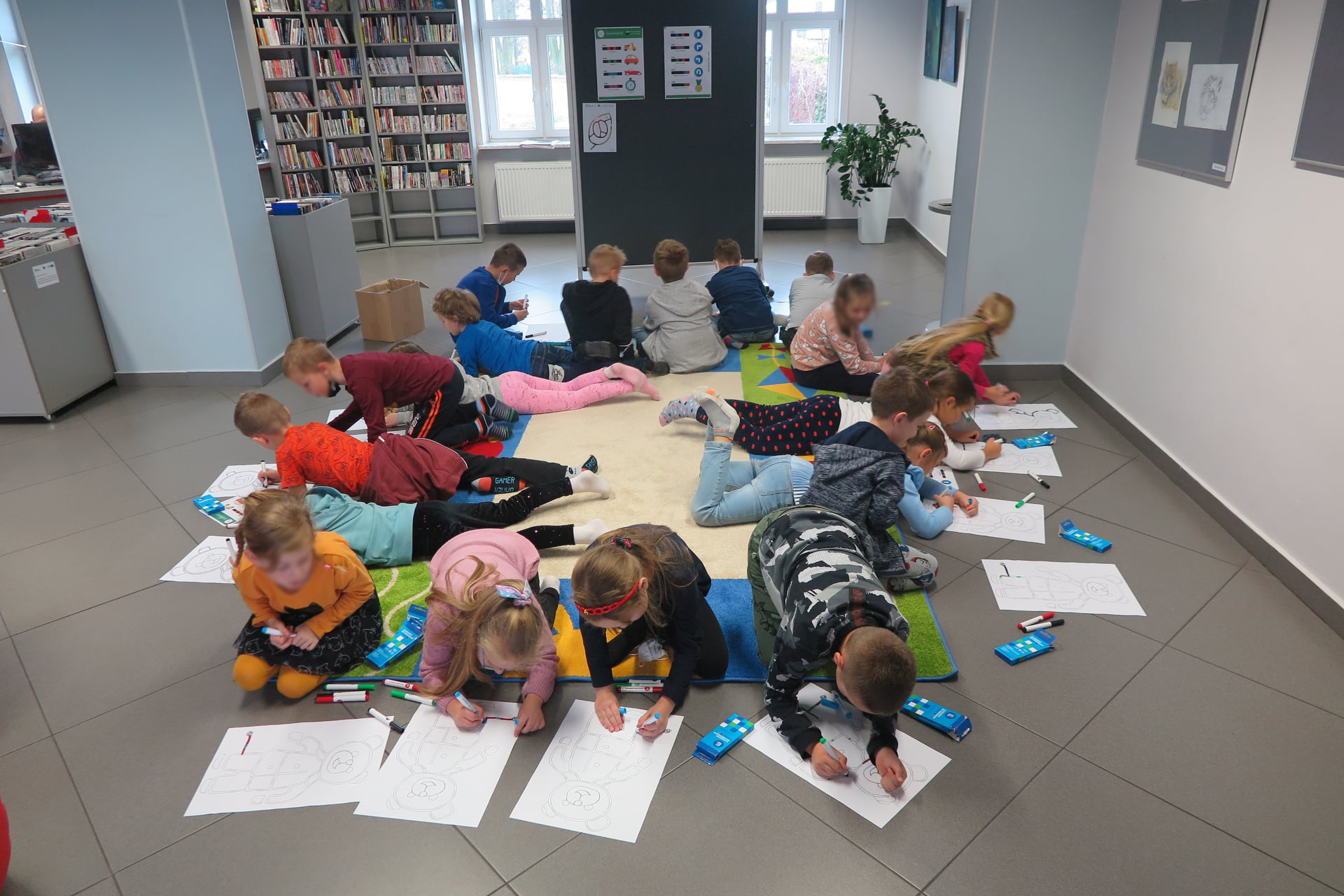 grupa dzieci siedzi/leży na dywanie skierowana na zewnątrz rysując flamastrami trasę dla osobotów w kształcie misia, w tle stoi tablica z przywieszonymi kodami ruchu ozobata