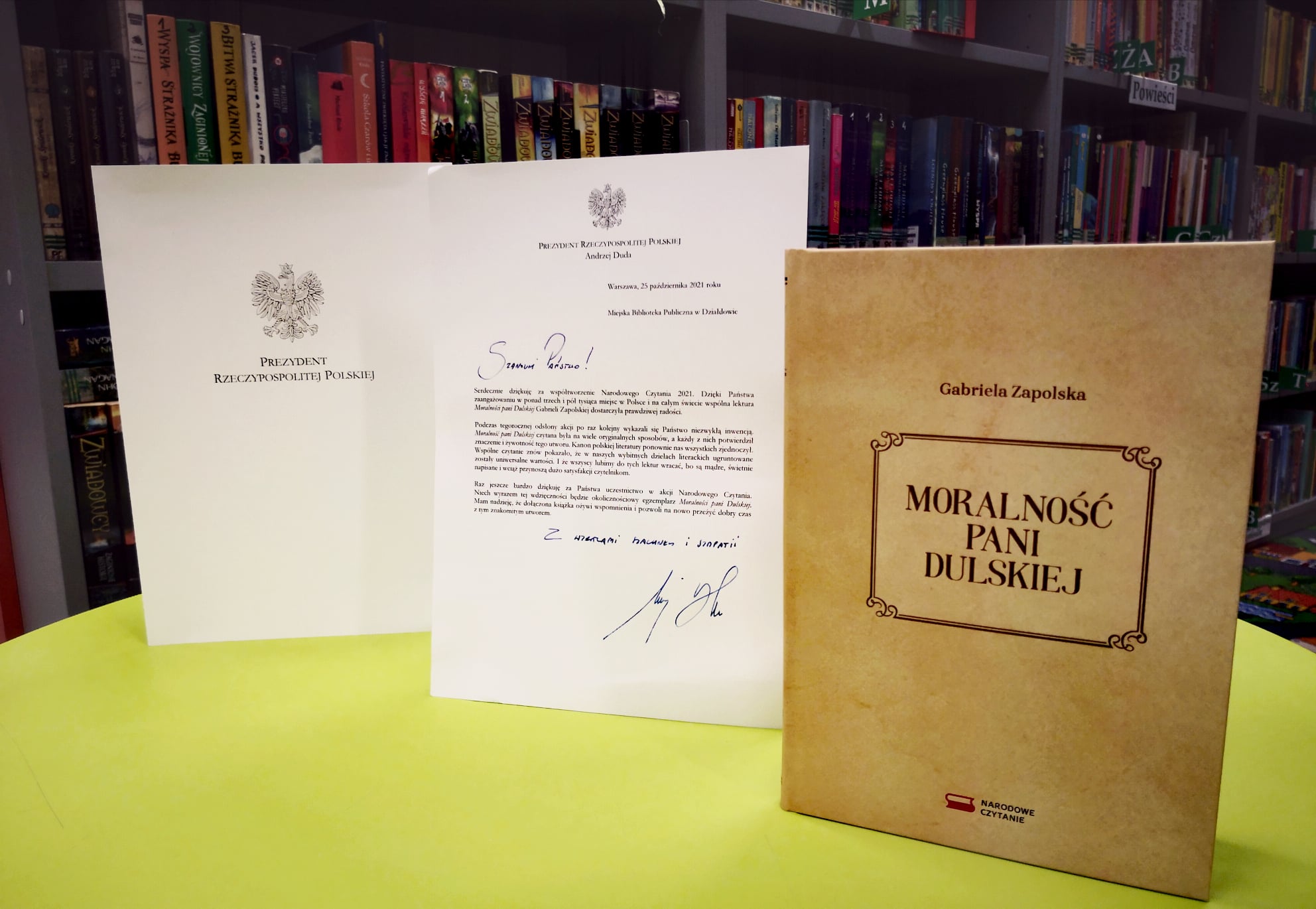 Wnętrze biblioteki, na stoliku stoi książka pt. "Moralność Pani Dulskiej" obok książki OBOK NIEJ LIST GRATULACYJNY OD Prezydenta Andrzeja Dudy.