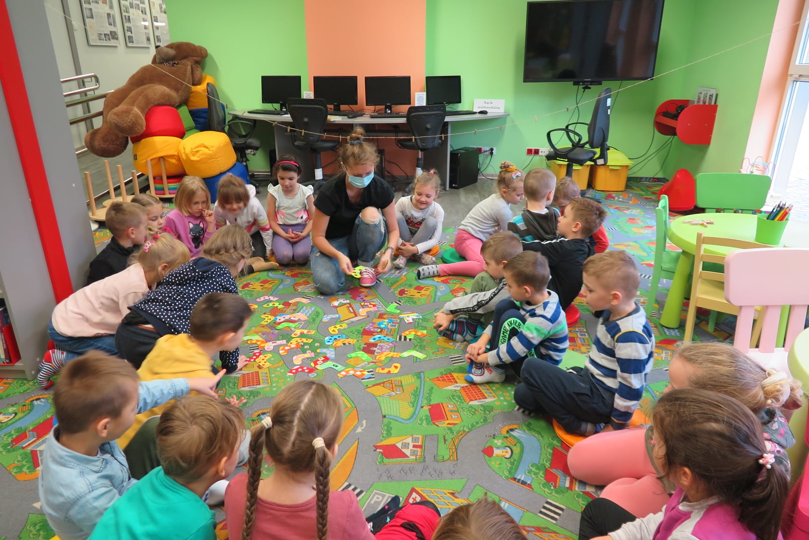 na dywanie w oddziale dla dzieci siedzi grupa przedszkolaków z Przedszkola nr 3, w środku siedzi bibliotekarka Aleksandra, przed dnia leżą kolorowe papierowe kolorowe skarpetki