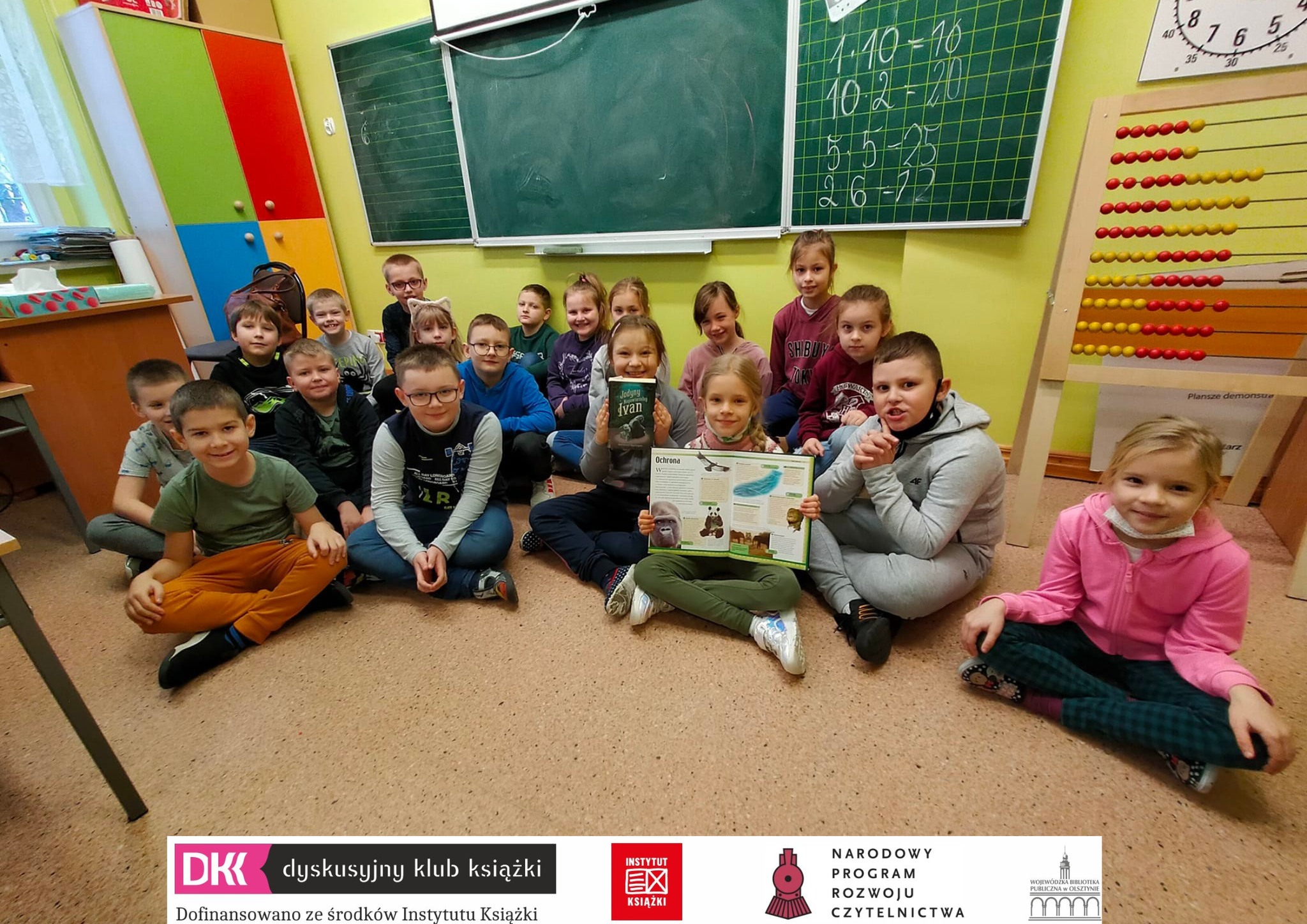 19 dzieci siedzi na podłodze pod tablicą w szkolnej klacie, dwie z nich trzymają książki