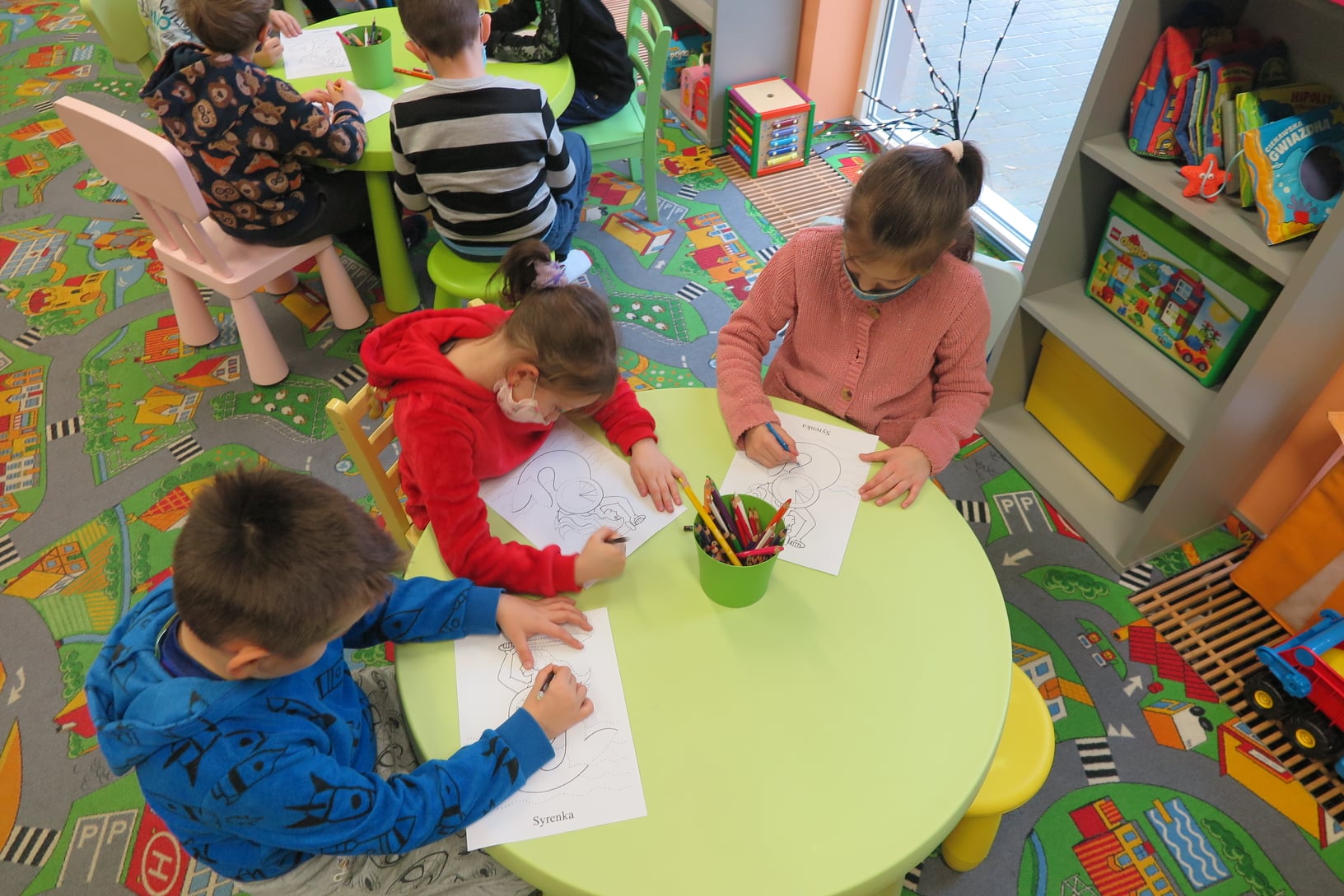 6 dzieci siedzi przy dwóch stolikach i koloruje kredkami herb warszawy - syrenę warszawską