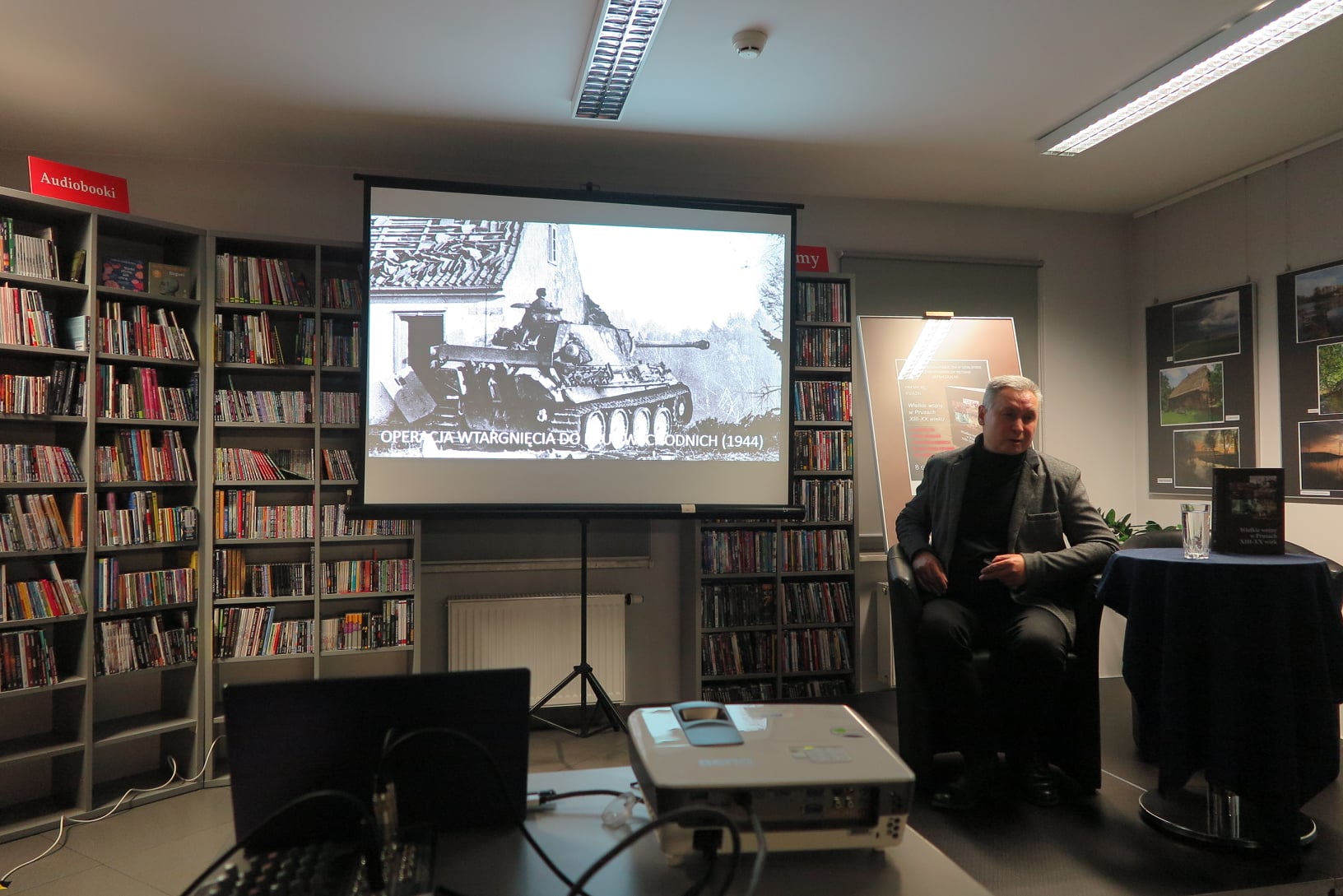 w fotelu siedzi dr Tomasz Gliniecki,  w tle plakat ze promocyjny i ekran projektora z wyświetlonym czarno=białym zdjęciem czołgu niemieckiego