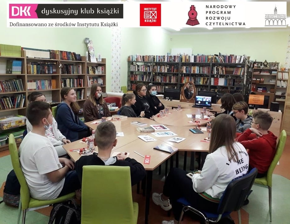 biblioteka szkolna, przy stole siedzi 13 uczniów i prowadzi spotkanie przez zooma z nauczycielką, na górze zdjęcia logo DKK