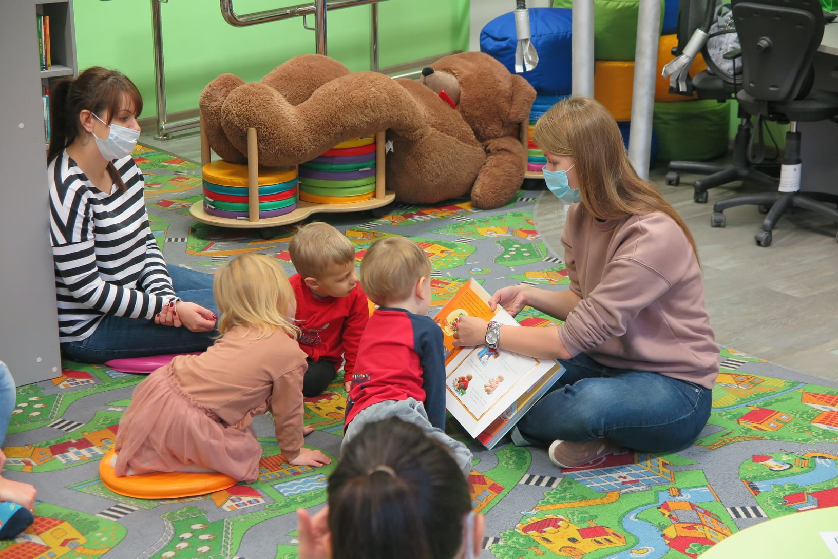wnętrze oddziału dla dzieci, na podłodze siedzą dzieci z rodzicami, Bibliotekarka Aleksandra pokazuje dzieciom obrazki w książce
