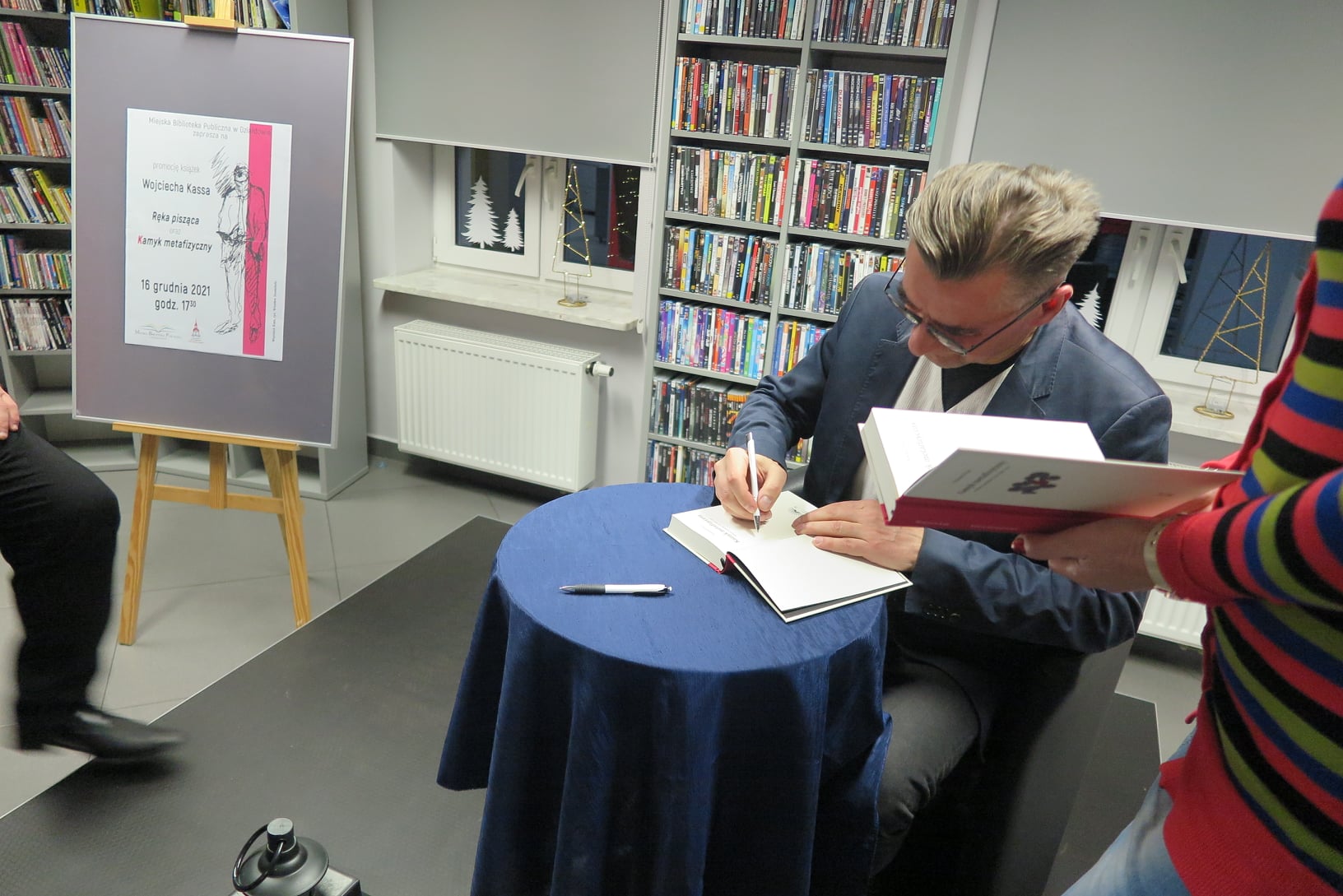 Wojciech Kass siedzi w fotelu przy okrągłym stoliku i podpisuje Publiczności egzemplarze książki