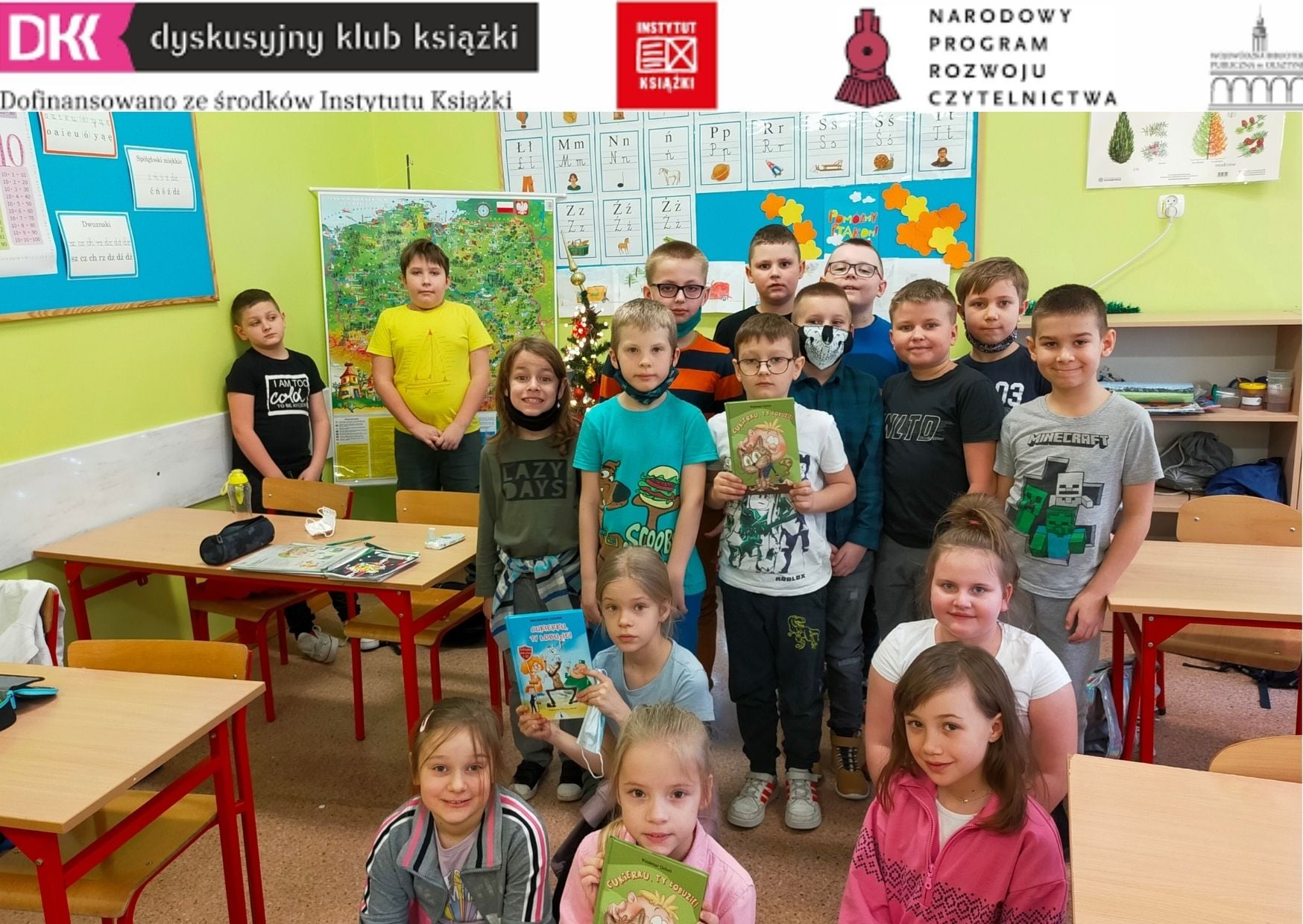 17 uczniów klasy 2b w sali lekcyjnej, troje z nich trzyma omawianą na spotkaniu DKK książką