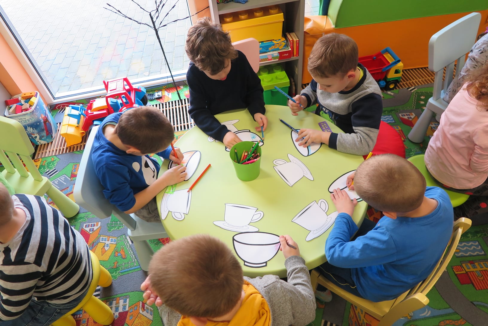 7 dzieci siedzi przy stoliku i koloruje wydrukowane filiżanki kawy