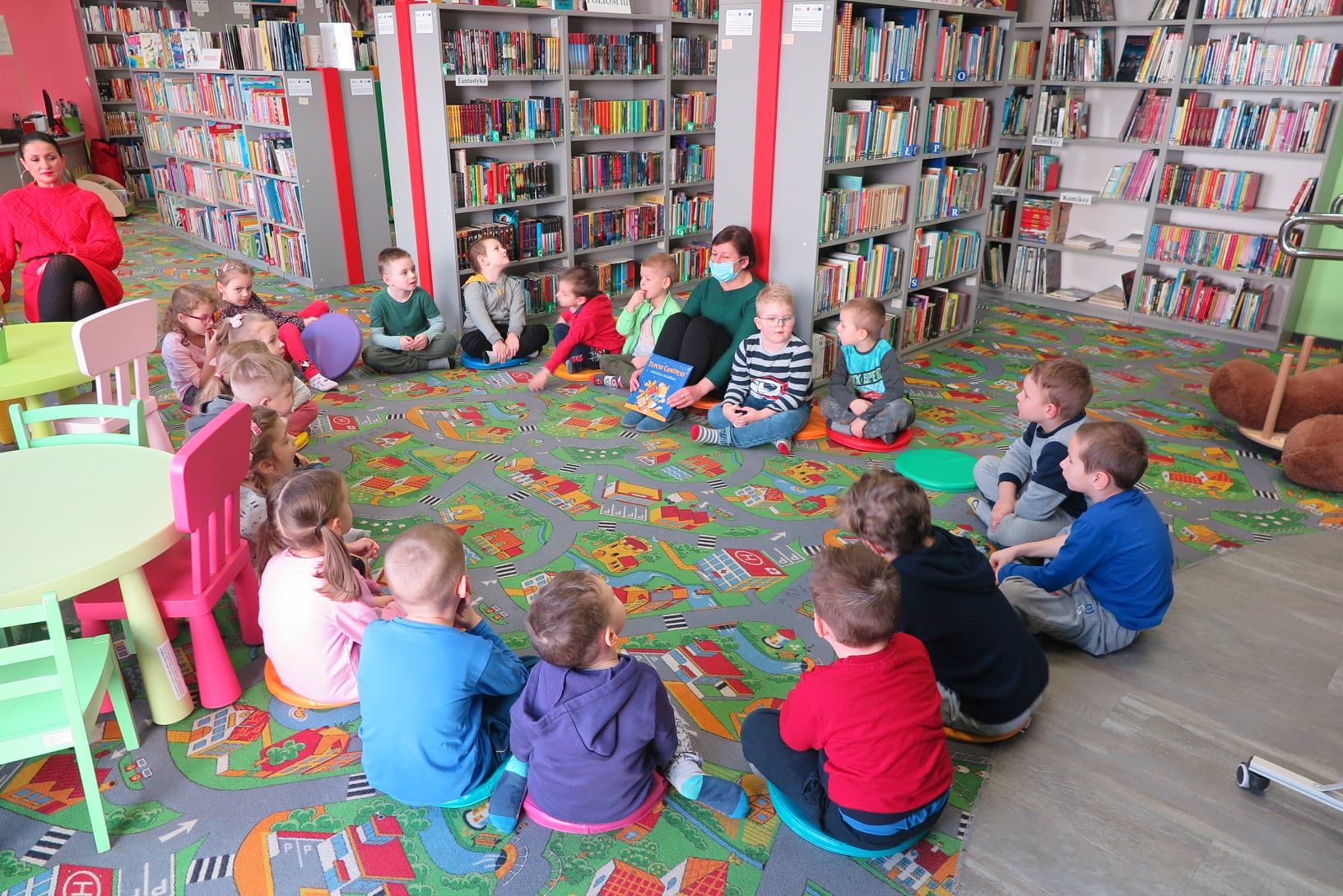 wnętrze oddziału dla dzieci biblioteki, dzieci siedzą w kołku z bibliotekarką Anią która trzyma w rękach książką. w tle regały z książkami