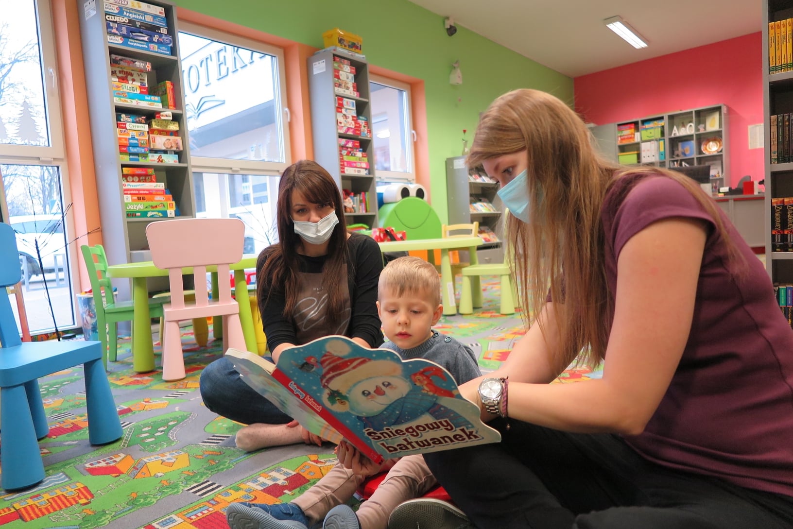 oddział dla dzieci, dwie kobiety i dziecko siedzą na dywanie, jedna trzyma w rękach otwartą książkę