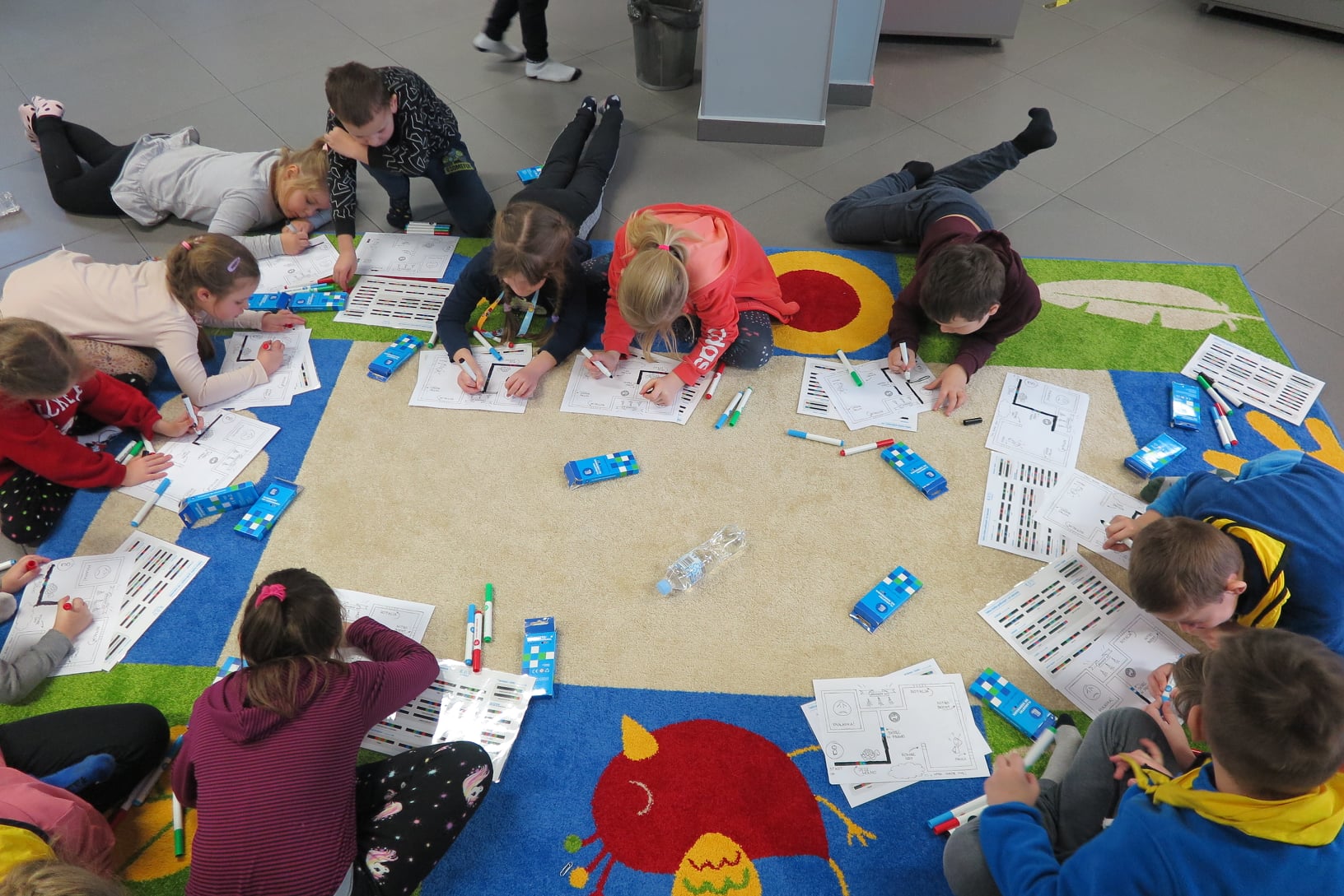 10 dzieci rysuje mazakami trasę na kartkach a4 dla ozobotów, dzieci siedzą na dywanie w mediatece