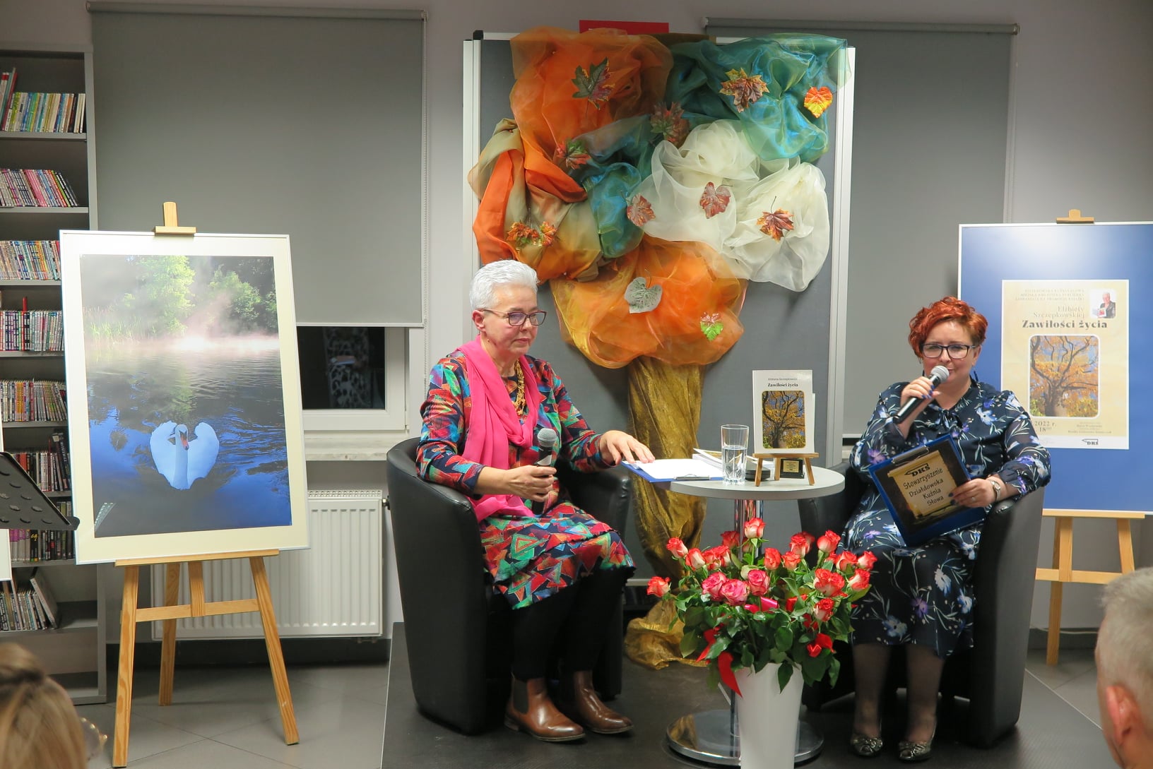 dwie kobiety siedzą na fotelach na podeście obie trzymają w rękach mikrofony, przed nimi na podłodze wazon z kwiatami, z tyłu po lewej zdjęci na sztaludze 