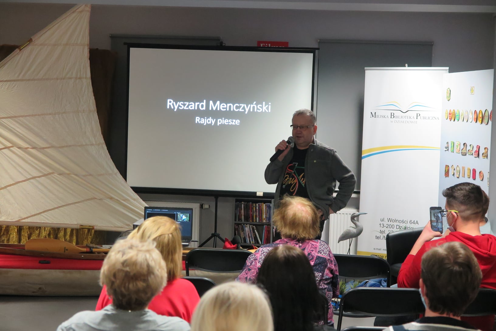 Ryszard Menczyński stoi z mikrofonem , w tle składany kajak z postawionym żaglem i ekran projektora, z przodu widać głowy publiczności