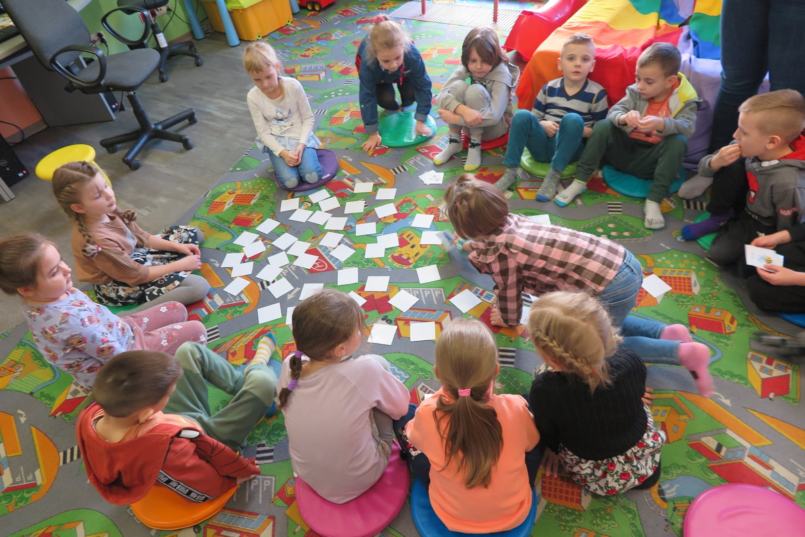 grupa dzieci siedzi w kółku na dywanie  i losuje kartki rozrzucone na podłodze
