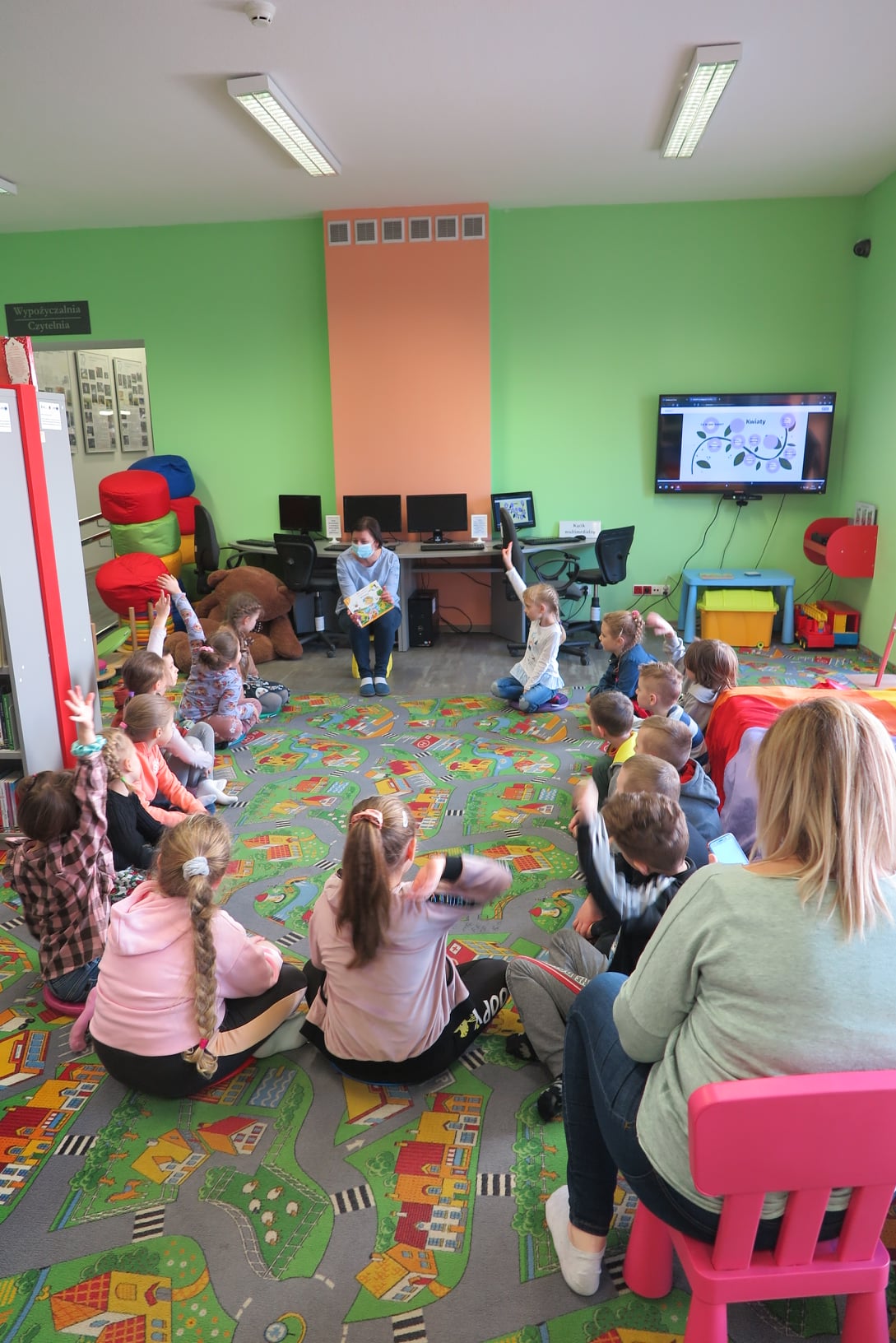 dzieci siedzą w kółko na podłodze z bibliotekarką trzymającą książkę , niektóre podnoszą ręce do góry