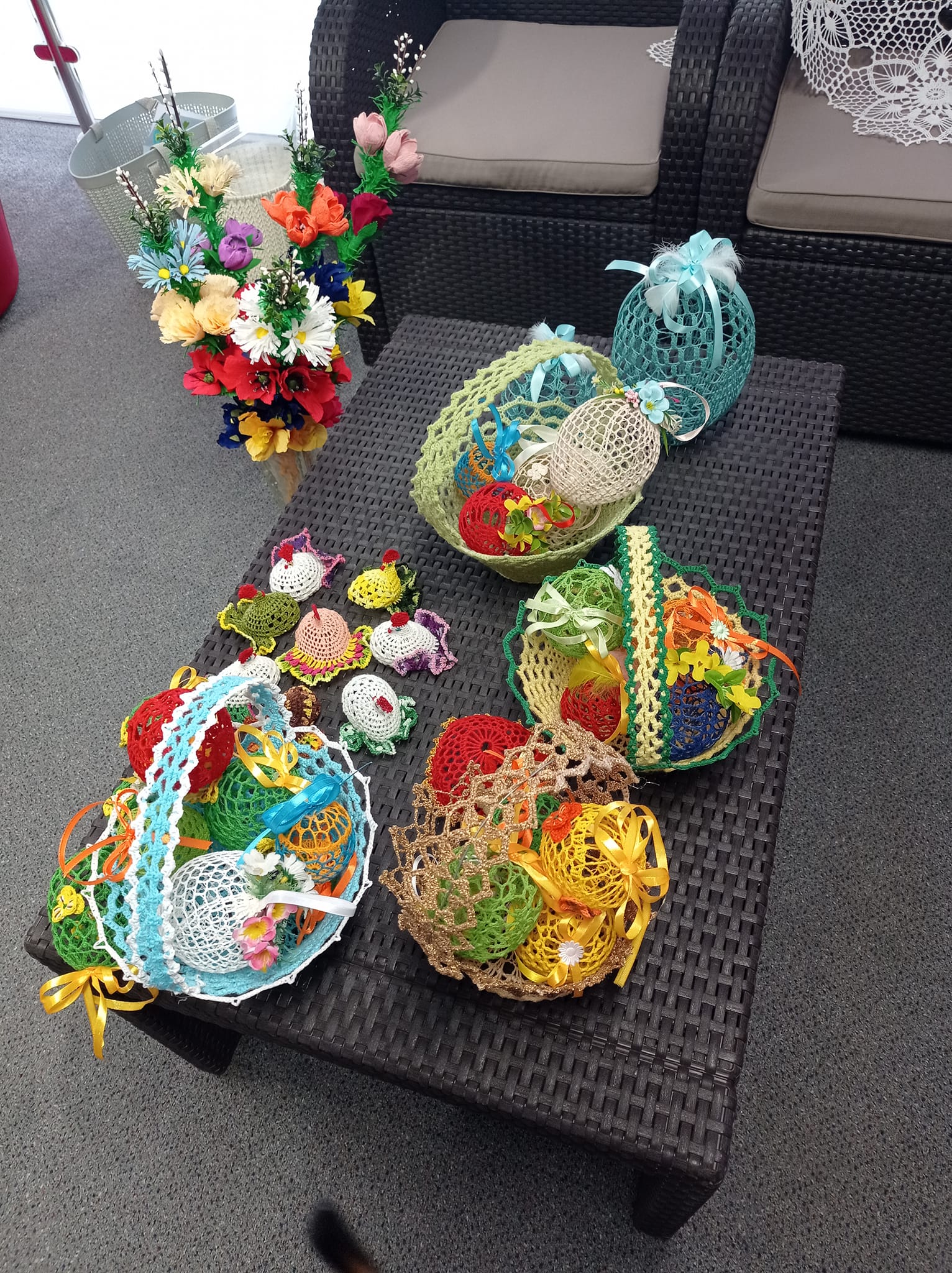 haftowany koszyki z haftowanymi jajkami i inne ozdoby wielkanocne leżą na ratanowym stoliku, obok stoi wazon z kwiatami z krepy