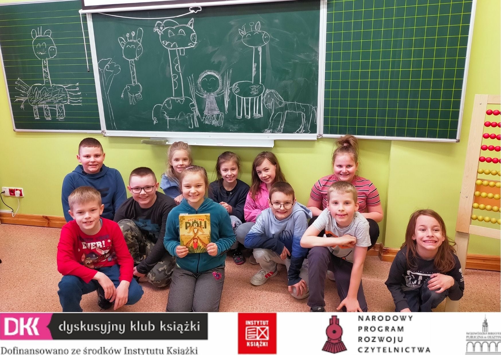 uczniowie klasy 2b w klacie siedzą pod tablicą , jedna z nich trzyma książką, na dole logo DKK i partnerów