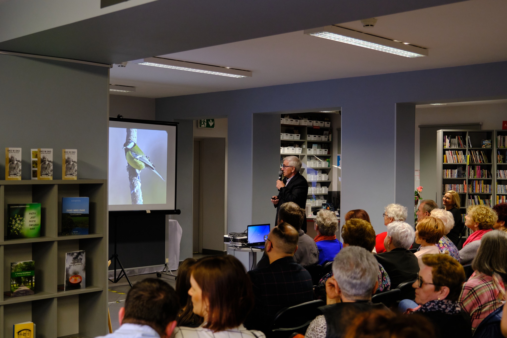 Ryszard Makszyński w garniturze trzyma przy ustach mikrofon, z przodu zdjęcie siedzie widownia, na ekranie projektora  widać ptaka na gałęzi