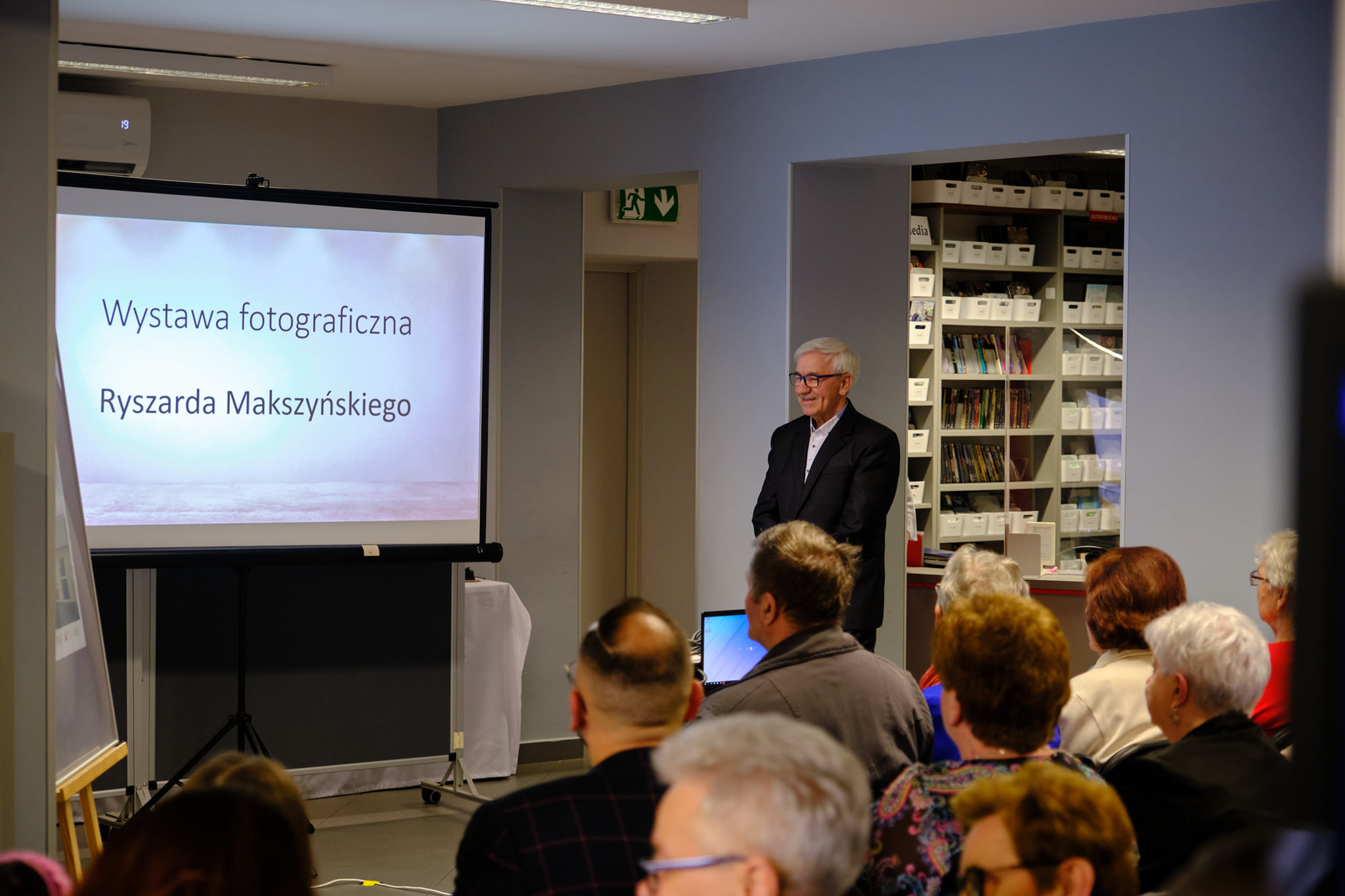 Ryszard Makszyński w garniturze stoi przy ekranie projektora, z przodu zdjęcie siedzie widownia, na ekranie projektora  widać napis wystawa fotograficzna ryszarda makszyńskiego