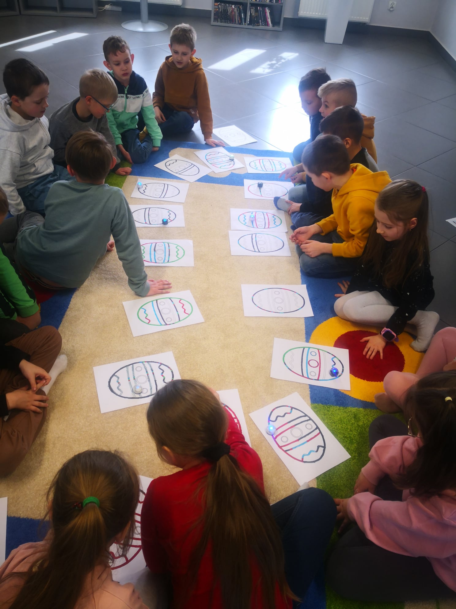 na dywanie w mediatece w kółku siedzi 15 dzieci, przed nimi leżą kartki a4 z narysowanymi trasami dla ozobota w kształcie wielkanocnej pisanki