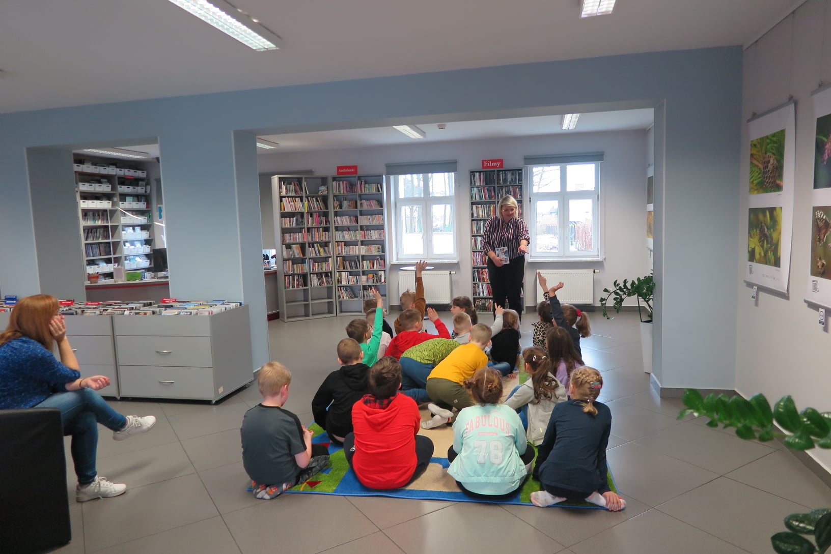 Mediateka, na środku sali rozłożony duży prostokątny dywan na którym siedzi 19 dzieci odwróconych plecami do fotografa. Przed dziećmi stoi bibliotekarka i prowadzi z nimi rozmowę pokazując coś dłonią.